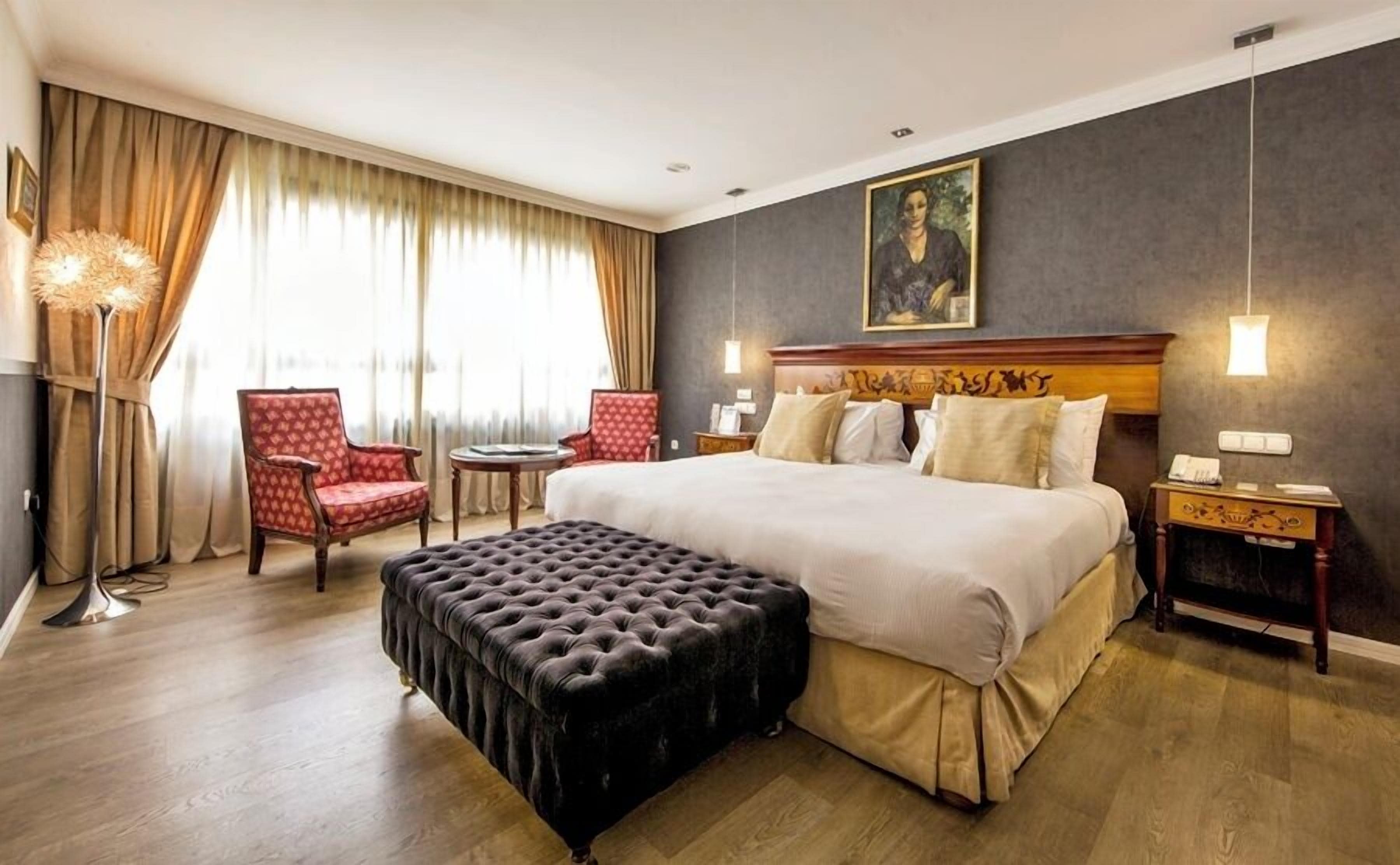 Une chambre d’hôtel avec un grand lit, deux fauteuils, une table ronde, un pouf capitonné et un tableau au mur.