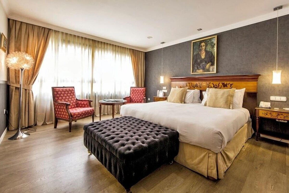 A hotel room with a large bed, two armchairs, a round table, a tufted ottoman, and a painting on the wall.
