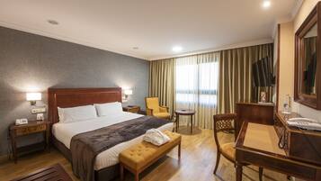Quarto de hotel com uma cama grande, mesas de cabeceira, escrivaninha, cadeira, pequena mesa e um espelho.