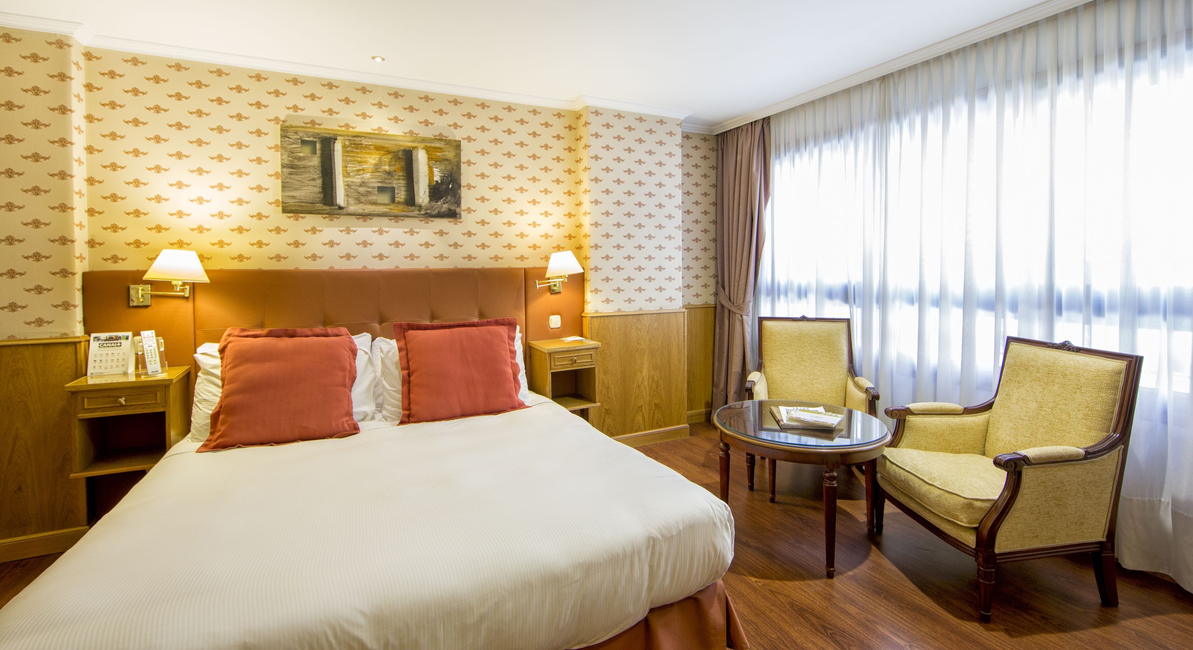 Ein Hotelzimmer mit einem großen Bett, zwei Nachttischlampen, einem kleinen Tisch, einem Sessel und einem großen Fenster mit Vorhängen.