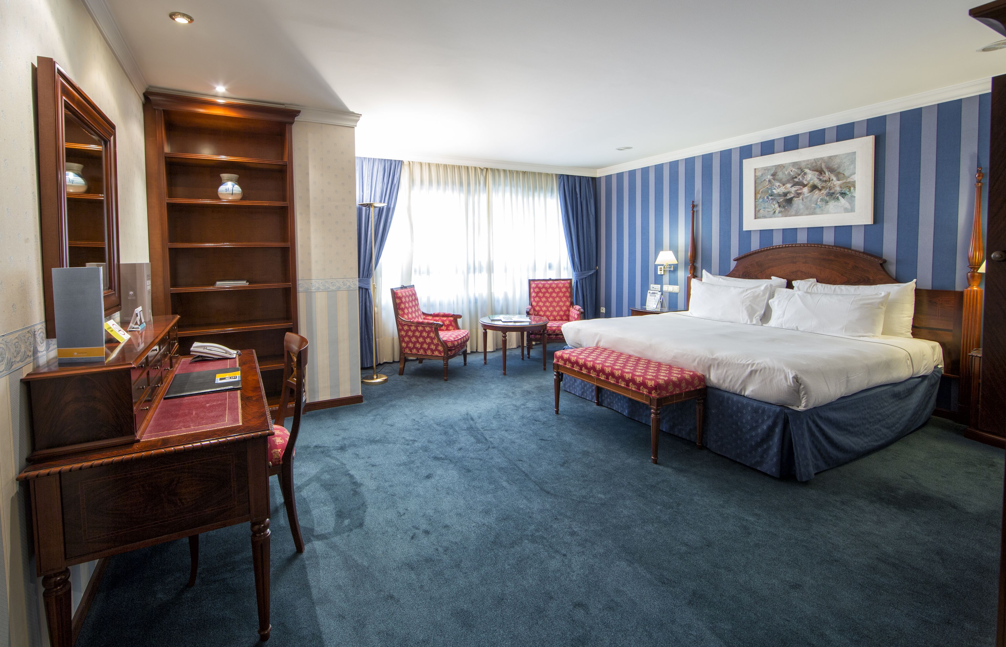 Habitación de hotel con una cama grande, un escritorio con una silla, una mesita con dos sillas, una estantería y una ventana con cortinas.