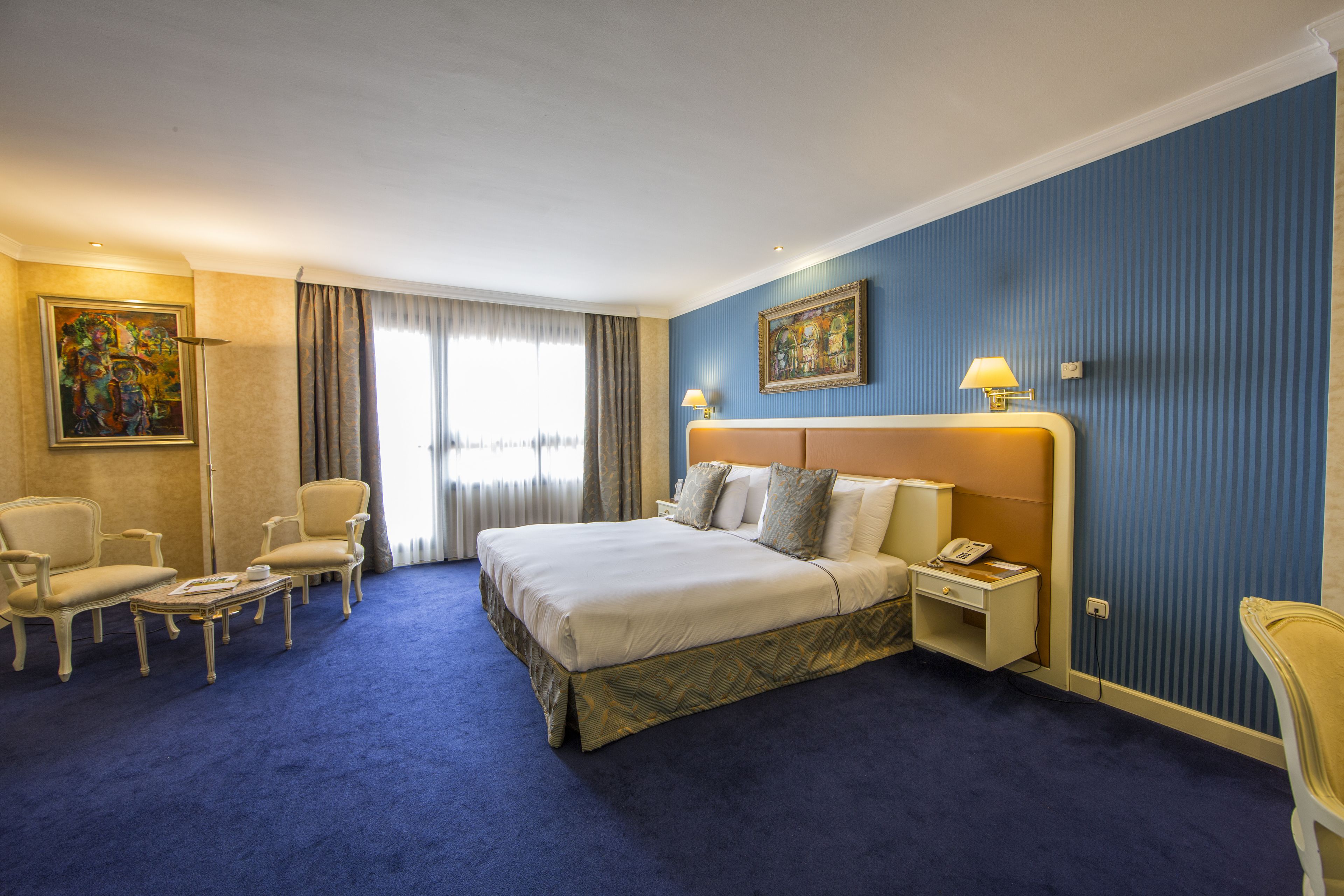 Habitación de hotel con una cama grande, dos sillones, una mesita, una mesita de noche con lámpara y un cuadro en la pared.