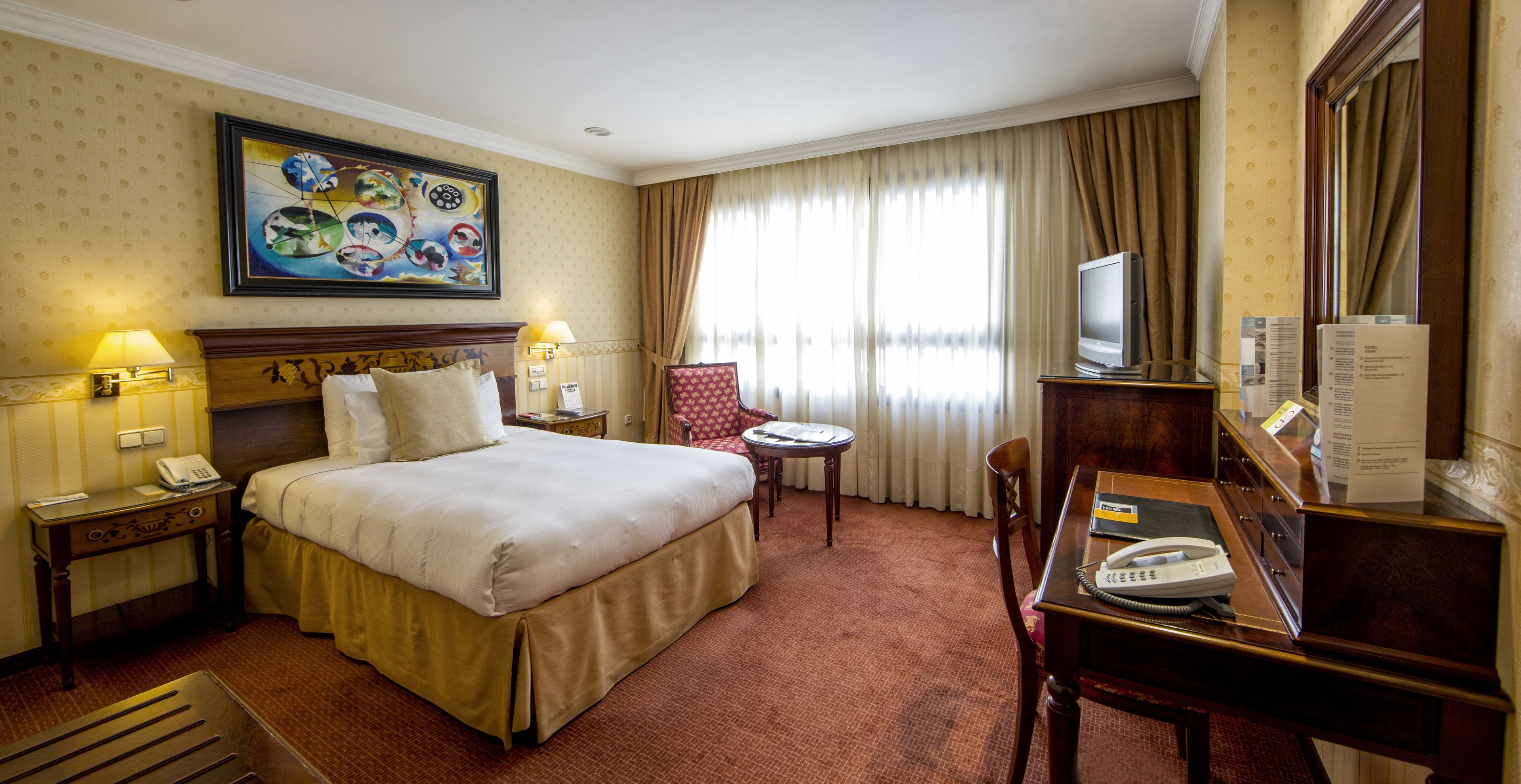 A hotel room with a large bed, a desk with a telephone, a chair, a small table, a TV, and a painting on the wall.