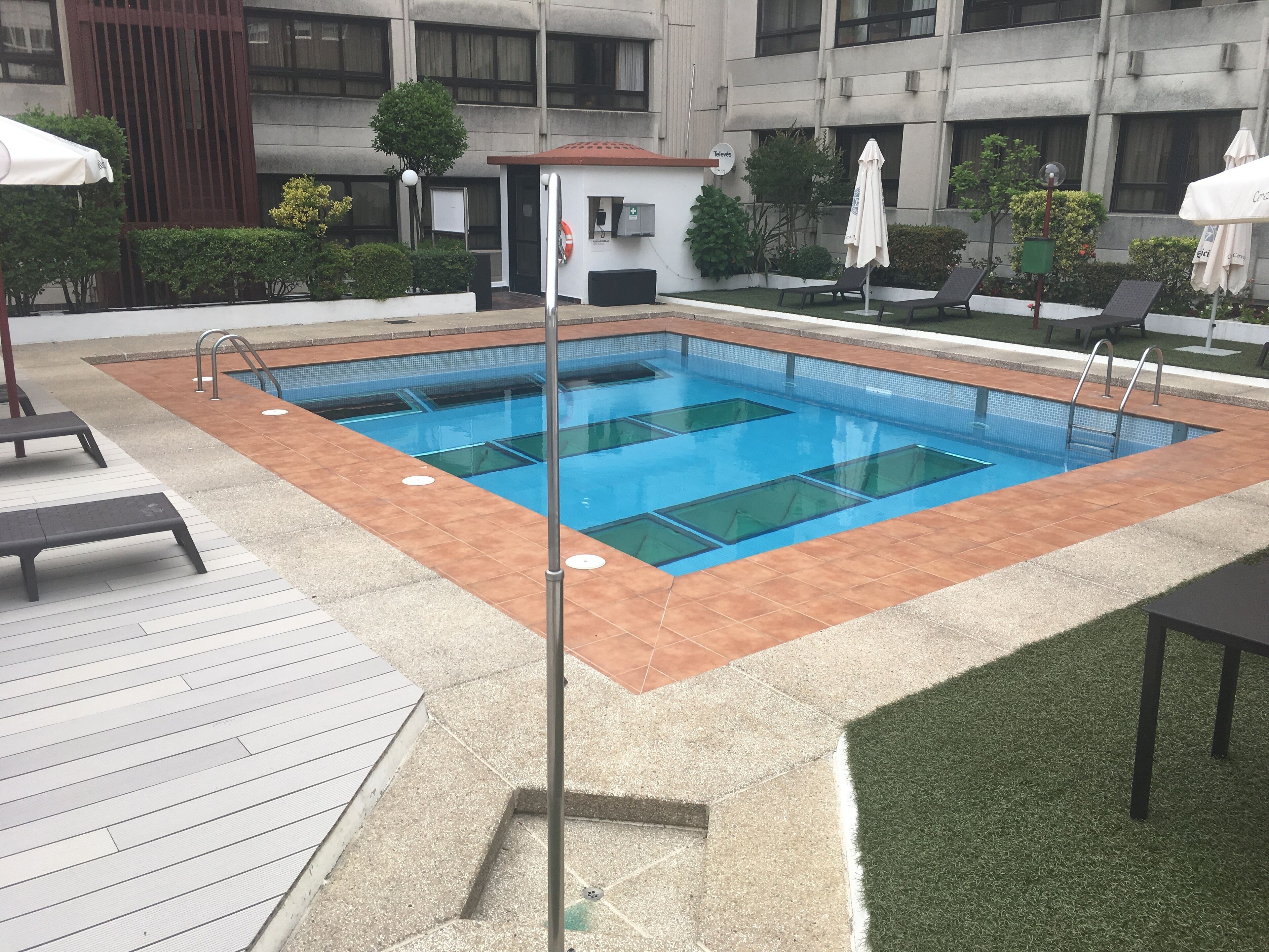 Una piscina rectangular con un borde empedrado, una zona cubierta con techo rojo y varias sillas de descanso y sombrillas.