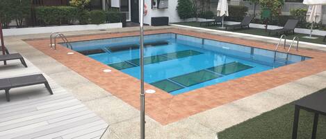 Une piscine rectangulaire entourée d’une terrasse carrelée, un petit espace couvert avec un toit rouge, ainsi que plusieurs chaises longues et parasols.