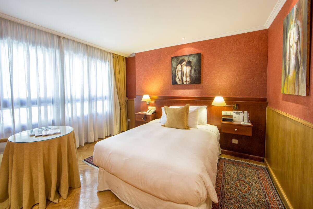 Een hotelkamer met een bed, nachtkastjes, een lamp, een schilderij aan de muur en uitzicht door het raam.