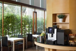 A modern cafe with a glass wall, a coffee machine, a plant, and a pastry display. - Somerset Park Suanplu Bangkok (Bangkok)