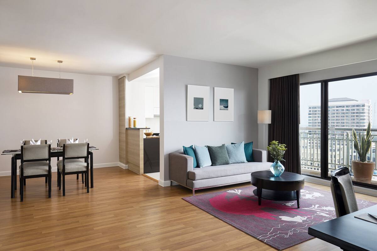 A modern living room with a dining area, a grey sofa with blue cushions, a round coffee table, and a large window with a city view.