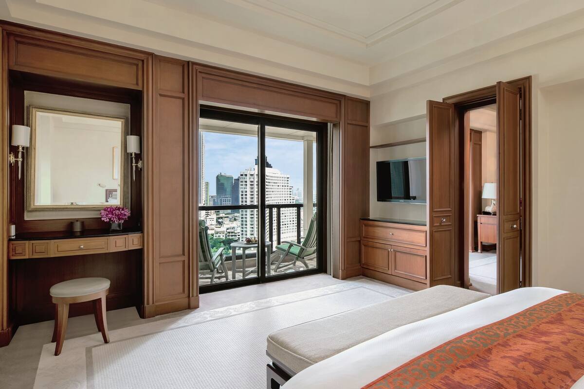 Ein Hotelzimmer mit einem großen Bett, einem Fernseher, einem Schreibtisch mit Stuhl, einem Schminktisch mit Spiegel und einem Balkon mit Blick auf die Stadt.