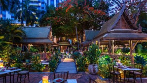 An outdoor dining area with tables set for two, surrounded by lush greenery and traditional wooden structures.