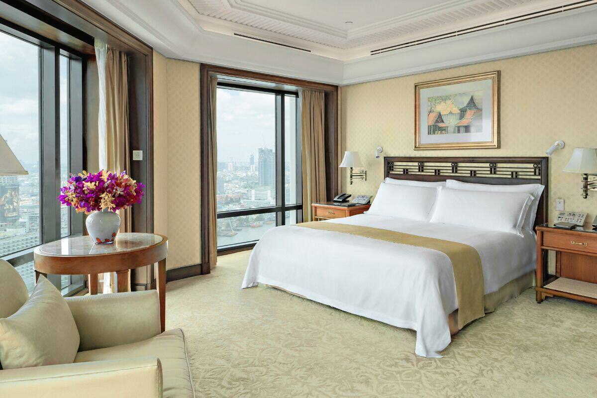 Ein Hotelzimmer mit einem großen Bett, einem runden Tisch mit Blumen in einer Vase, einem Sessel, einem Nachttisch mit einer Lampe und Blick auf die Stadt.