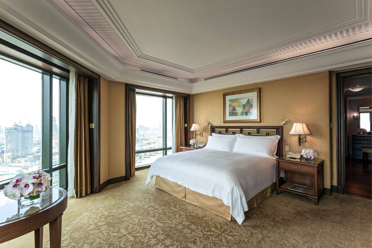 Ein Hotelzimmer mit einem großen Bett, zwei Nachttischen mit Lampen, einem runden Tisch mit Blumenvase und einem großen Fenster mit Blick auf die Stadt.