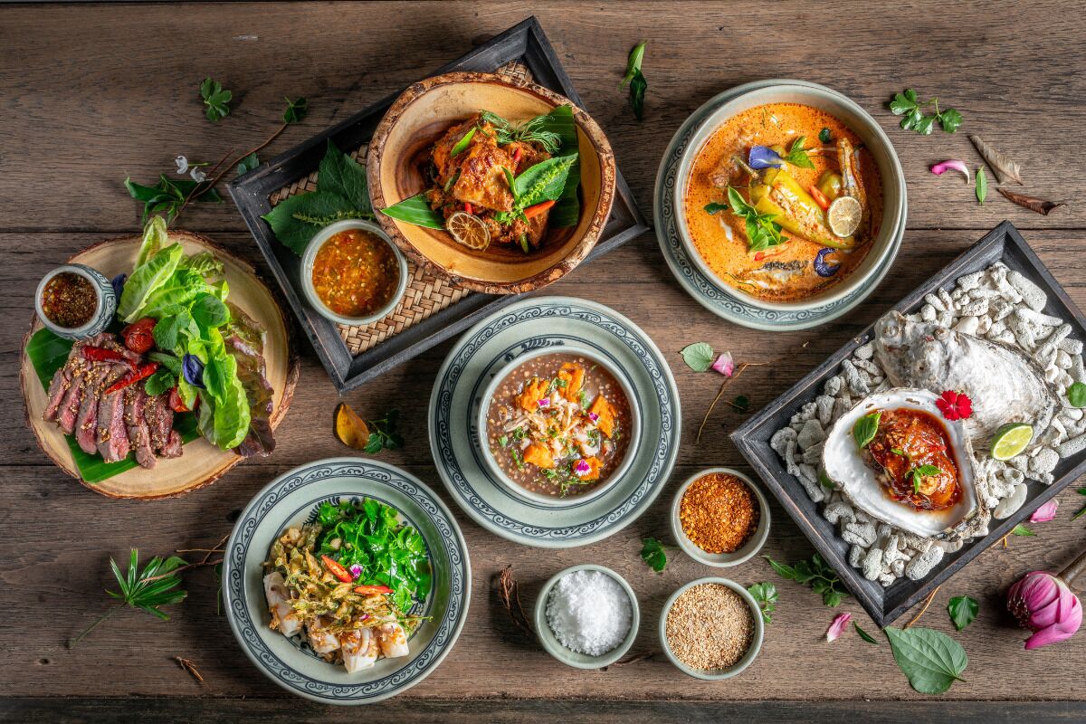 A variety of dishes including seafood, salads, and bowls of soup, arranged on a wooden table with various condiments and herbs.