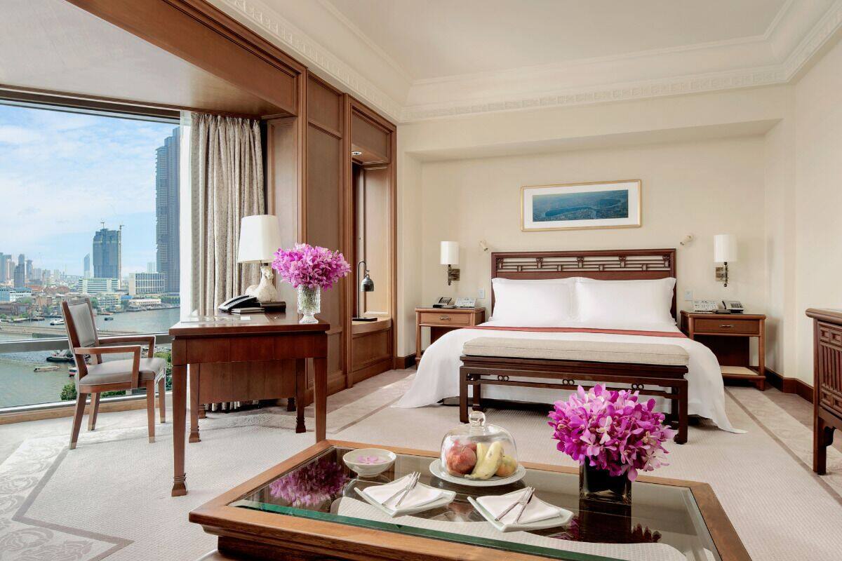 Ein Hotelzimmer mit einem großen Bett, einem Schreibtisch mit Blumen in einer Vase, Blick auf die Stadt und einem Esstisch mit Obstschale.