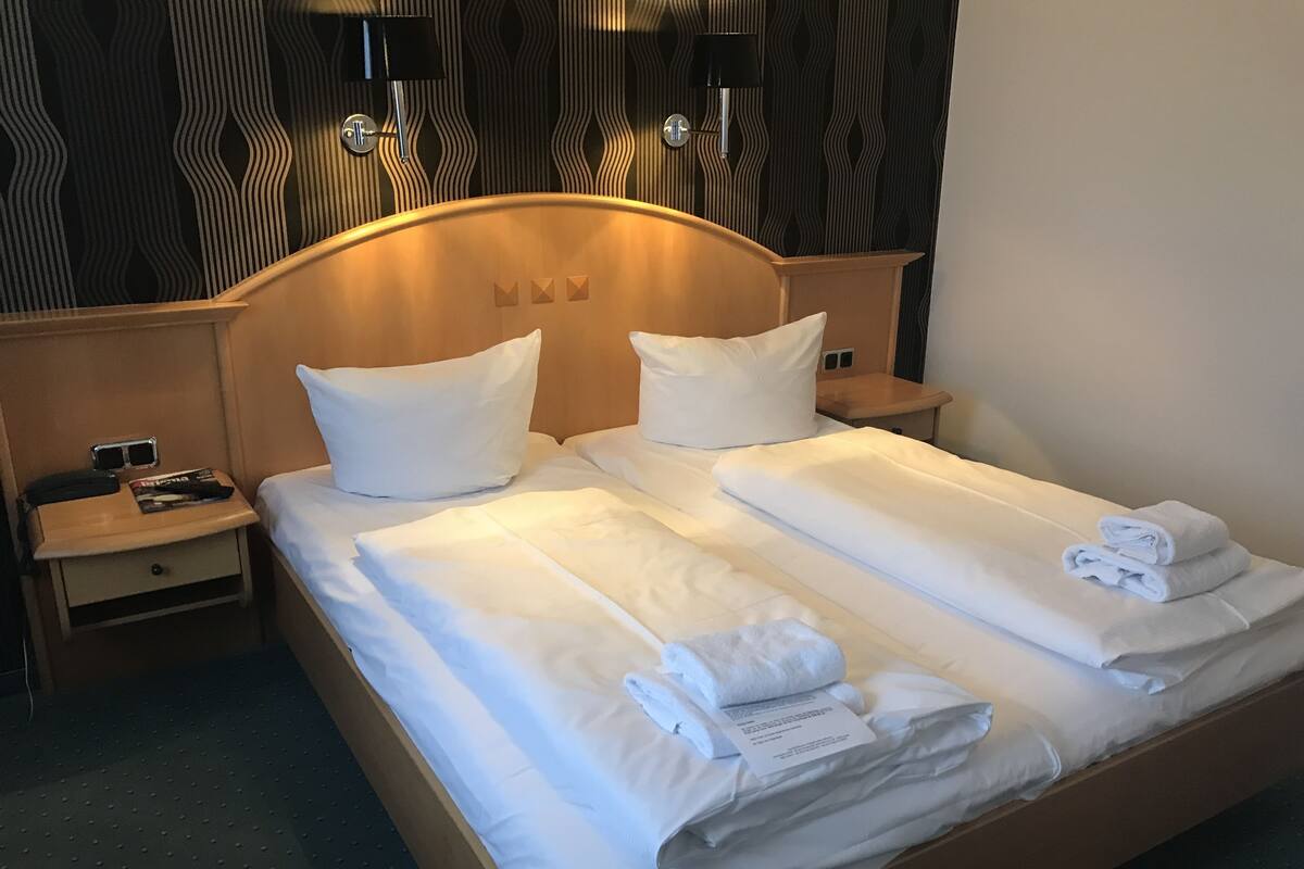 A double bed with white linens, two pillows, a folded towel, and a bedside table with a phone and a magazine.