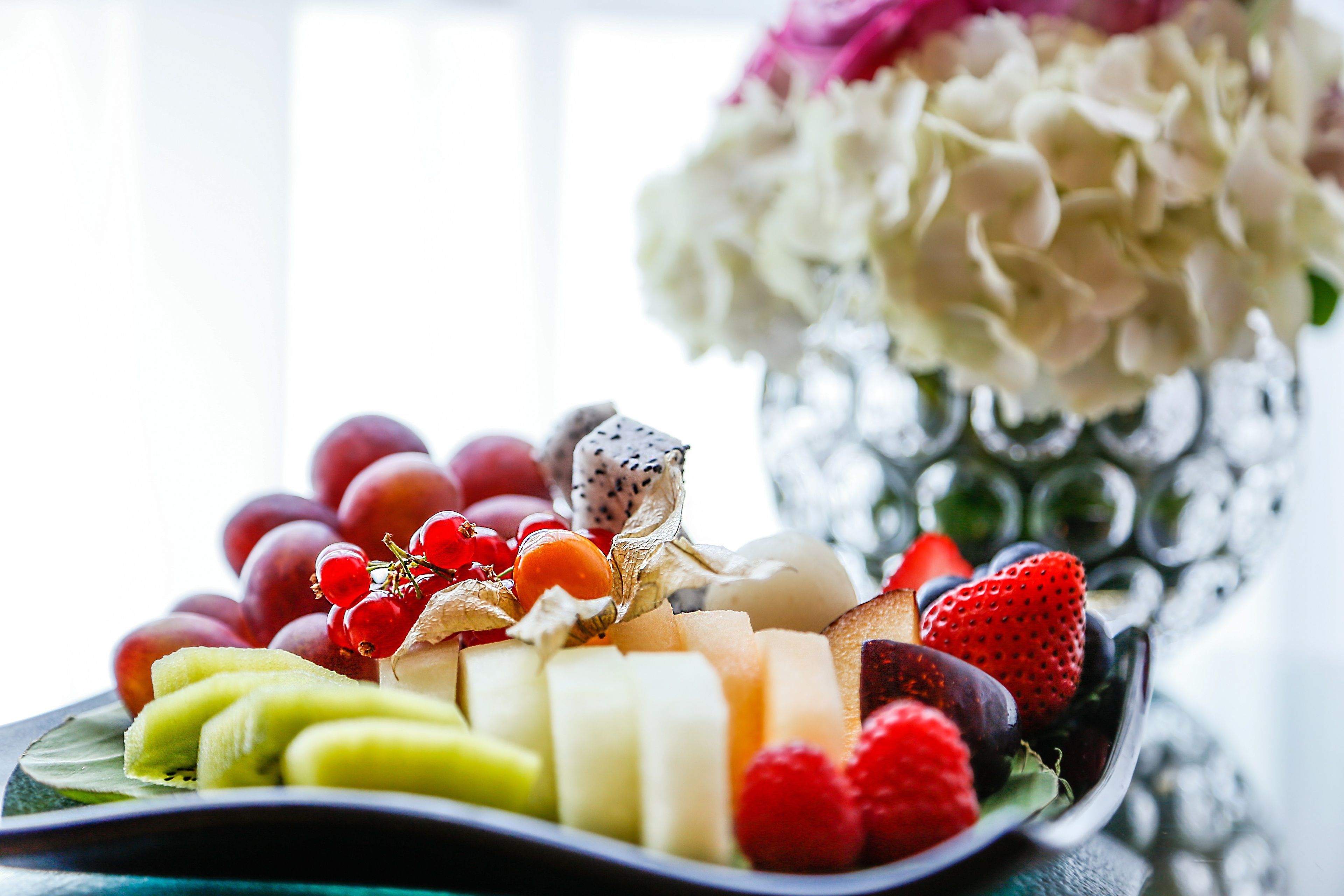 A fruit platter with various fruits and a decorative vase in the background.