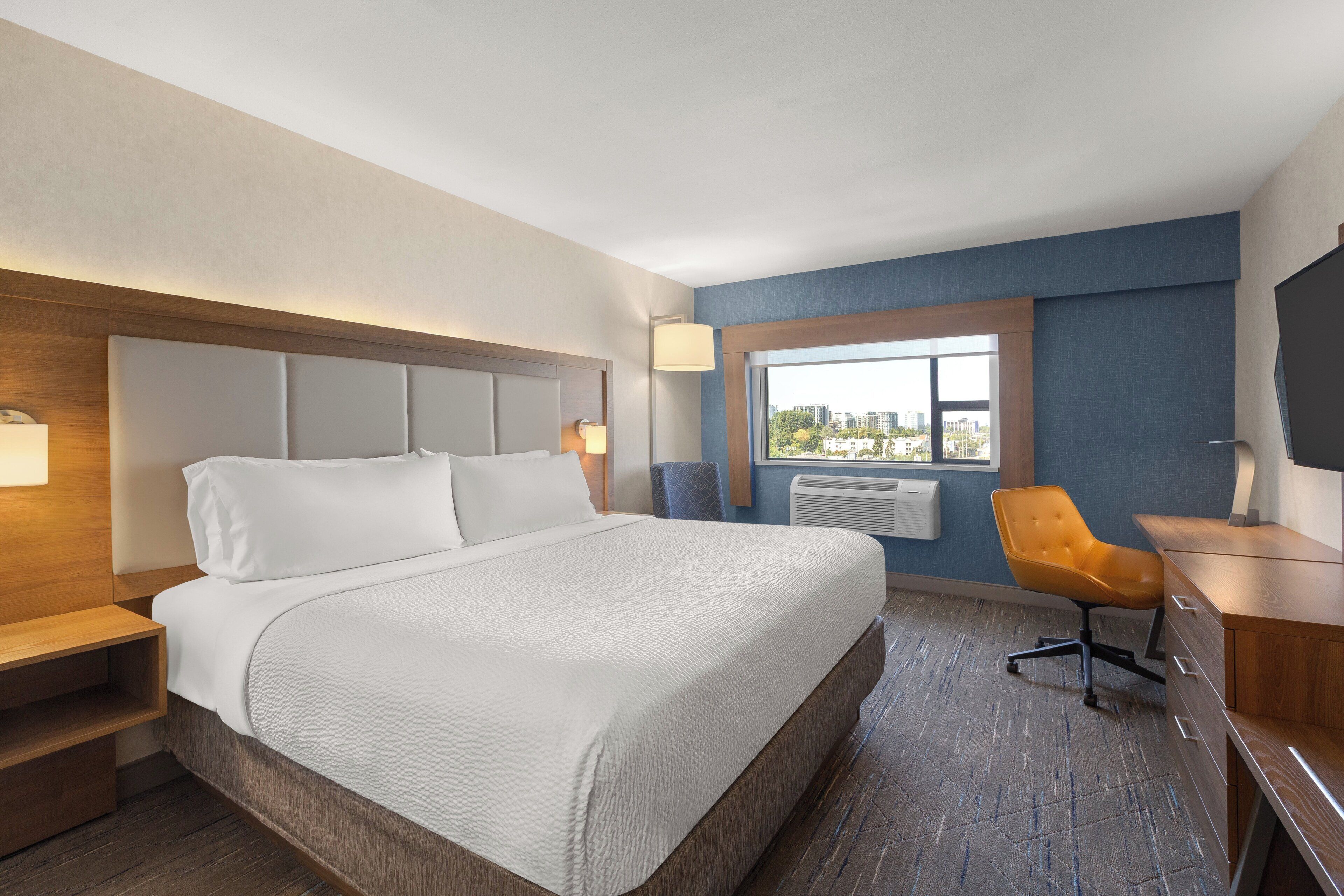 A hotel room with a large bed, a desk, a chair, and a window with a city view.