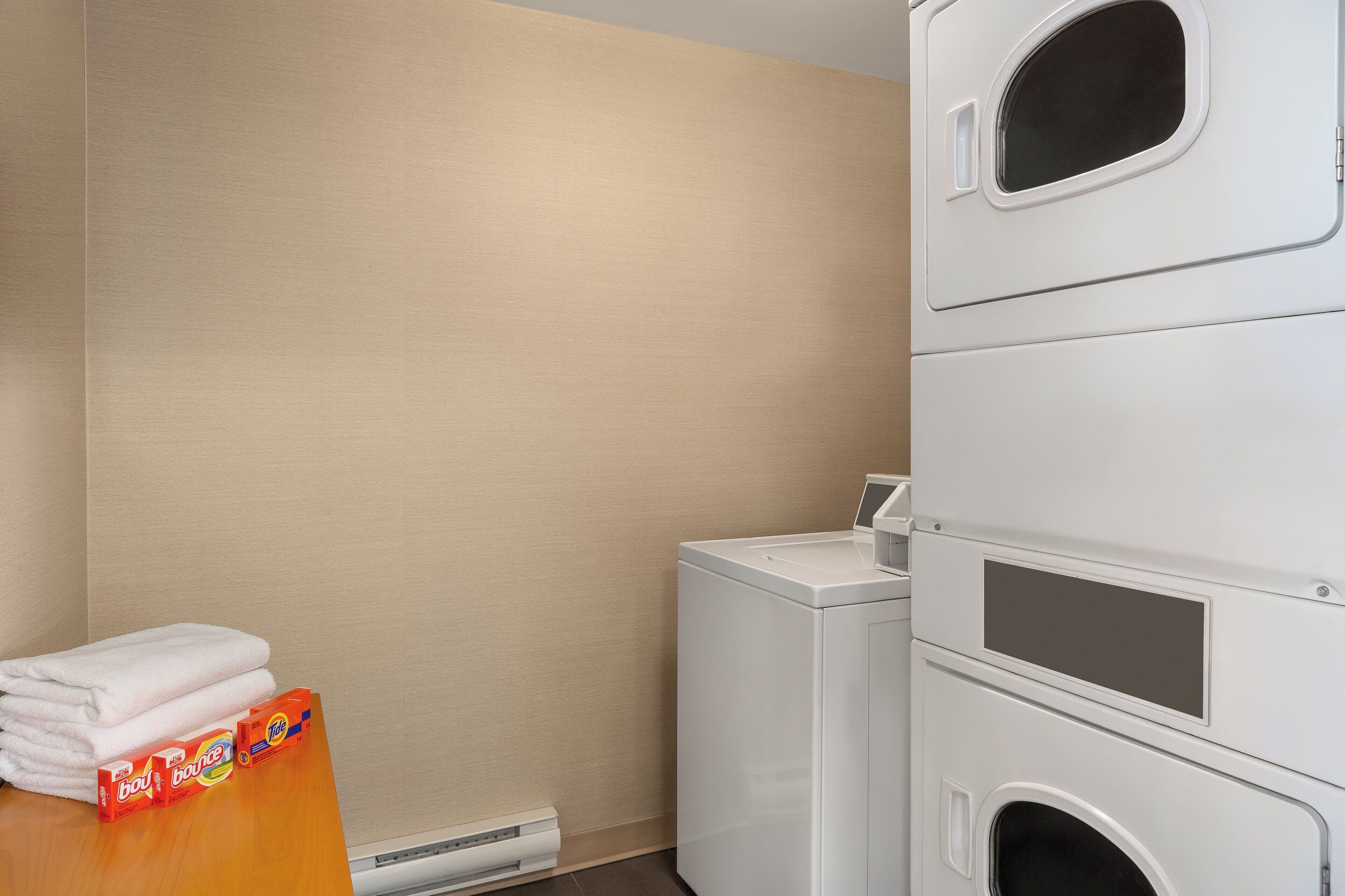 A laundry area with stacked white washing machines, a dryer, and a stack of white towels on a wooden counter.