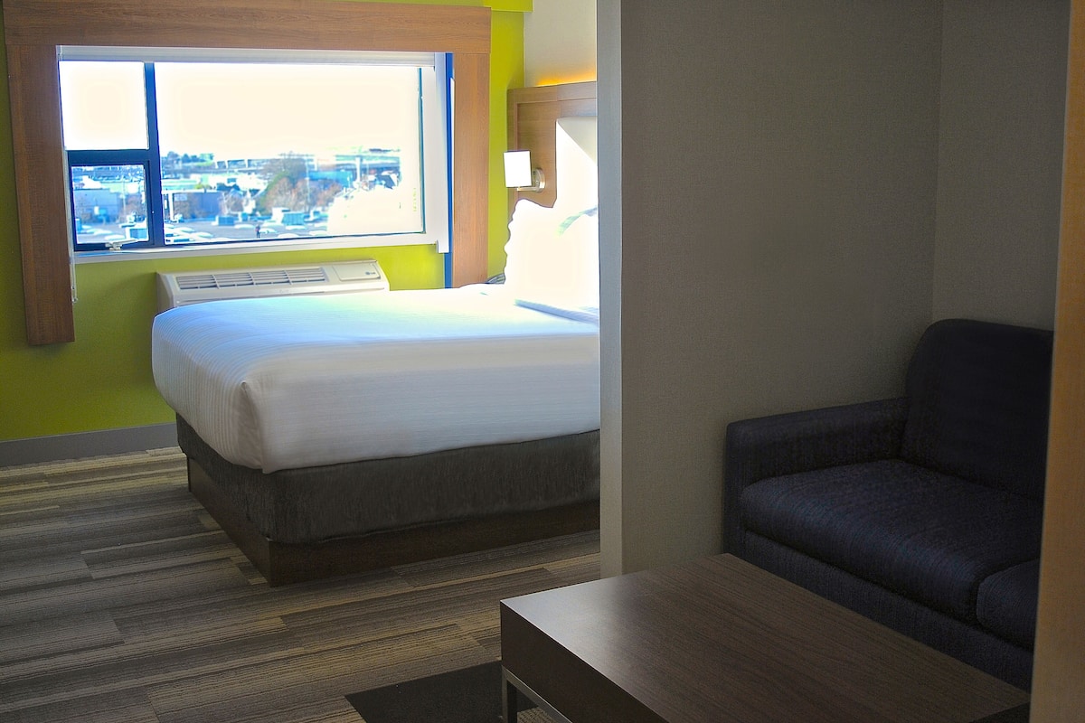 Une chambre d’hôtel avec un lit, un canapé, une fenêtre donnant sur des immeubles et un climatiseur fixé au mur.