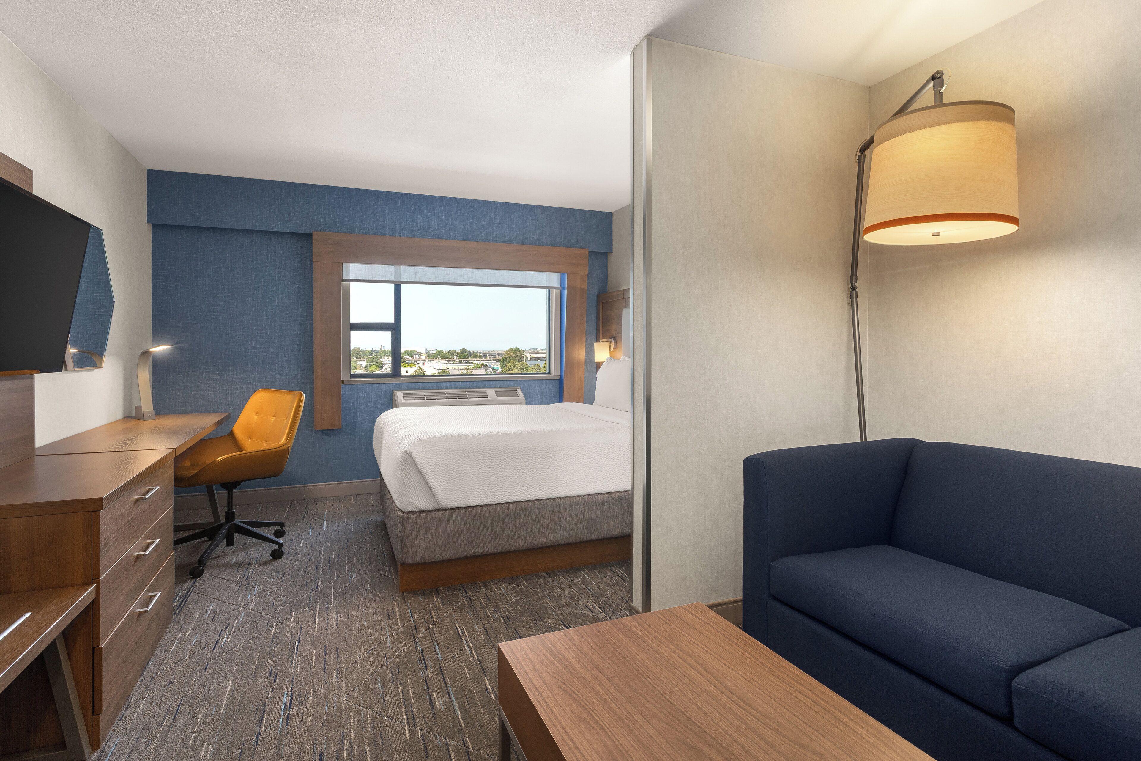 A hotel room with a bed, a desk, a chair, a sofa, a lamp, and a window with a view.