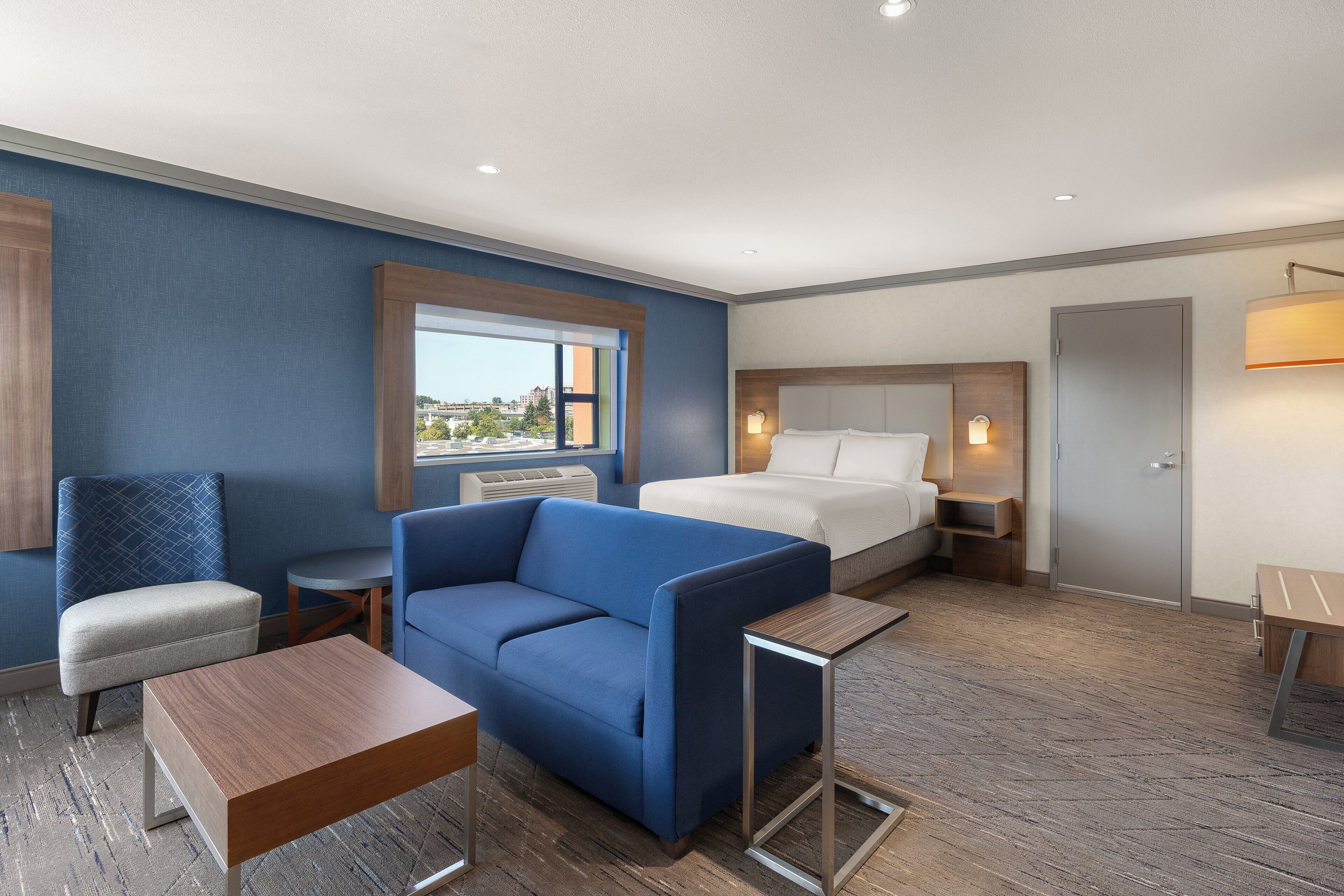 Habitación de hotel con cama, sofá azul, una mesita y ventana con vista a edificios.