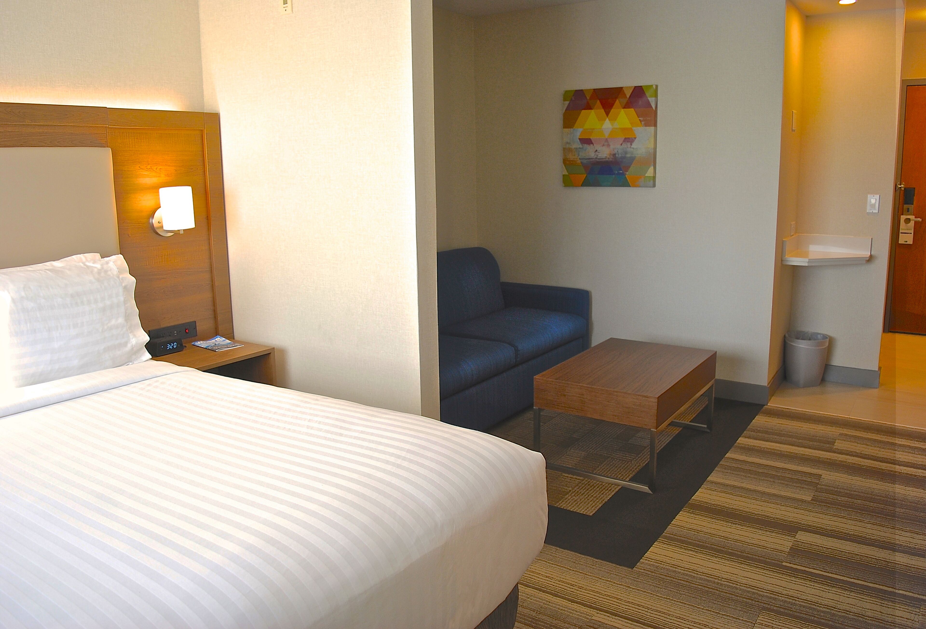 Una habitación de hotel con una cama grande, una mesita de noche, un sillón azul, una mesa de centro de madera y un cuadro en la pared.