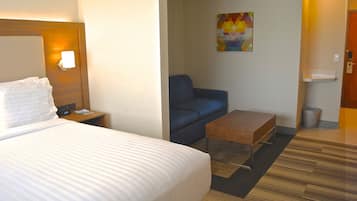 Una habitación de hotel con una cama grande, una mesita de noche, un sillón azul, una mesa de centro de madera y un cuadro en la pared.