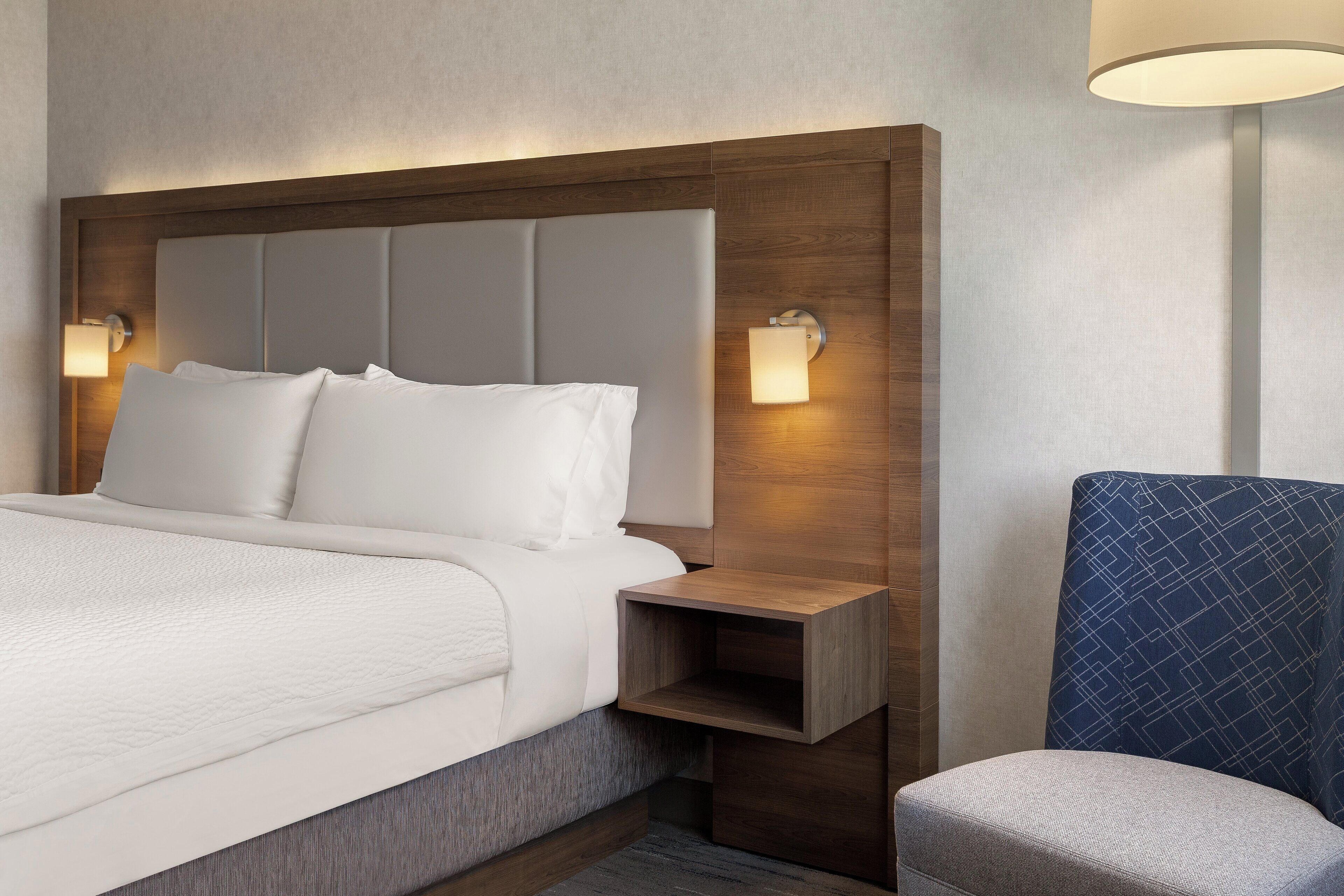 Quarto de hotel moderno com uma cama grande, cabeceira de madeira, duas luminárias de cabeceira e uma poltrona azul estofada.