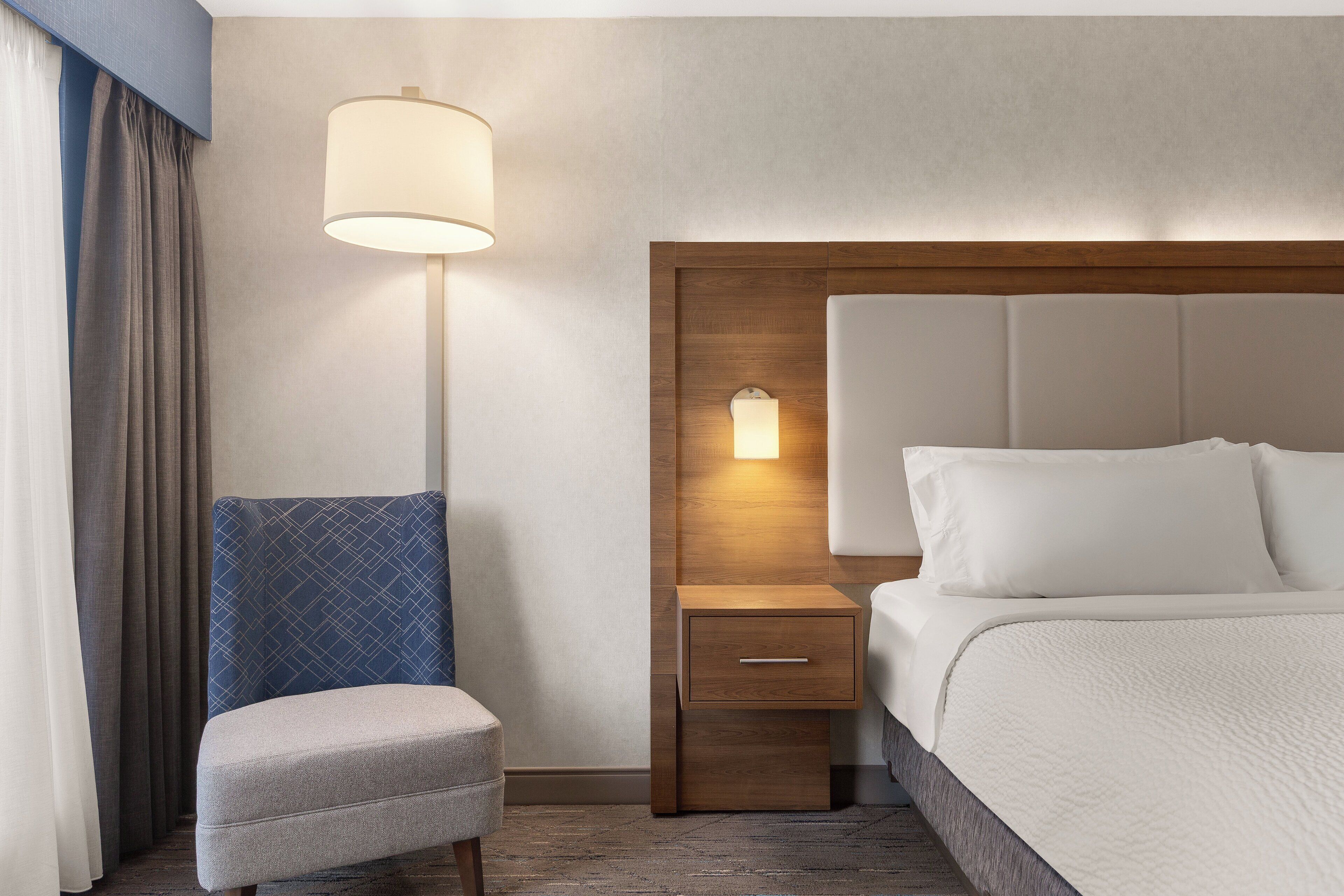 Une chambre d’hôtel avec un lit, une table de chevet avec une lampe, une chaise et une tête de lit en bois.