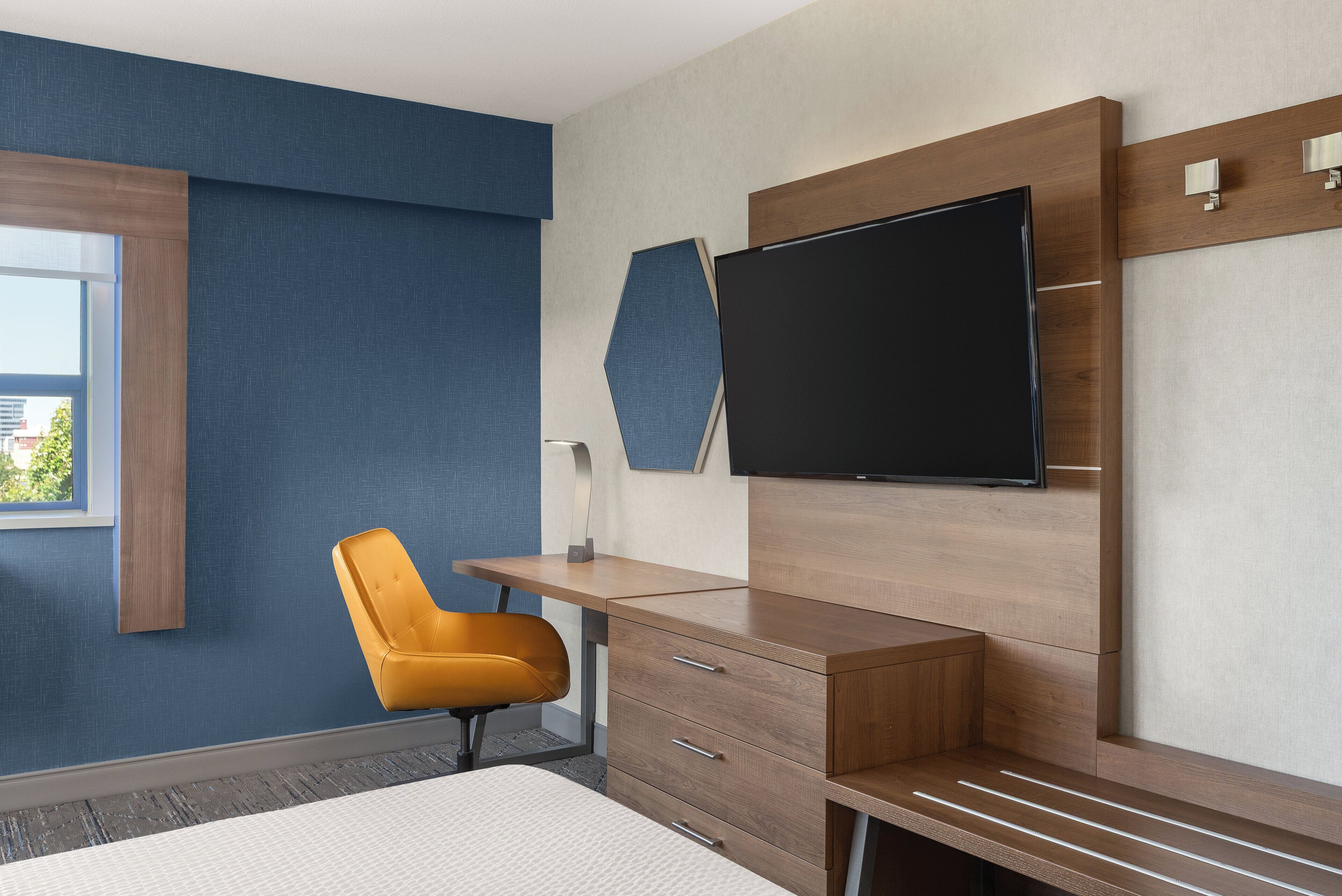A modern hotel room with a wooden desk, a flat-screen TV, a comfortable chair, and a large bed.
