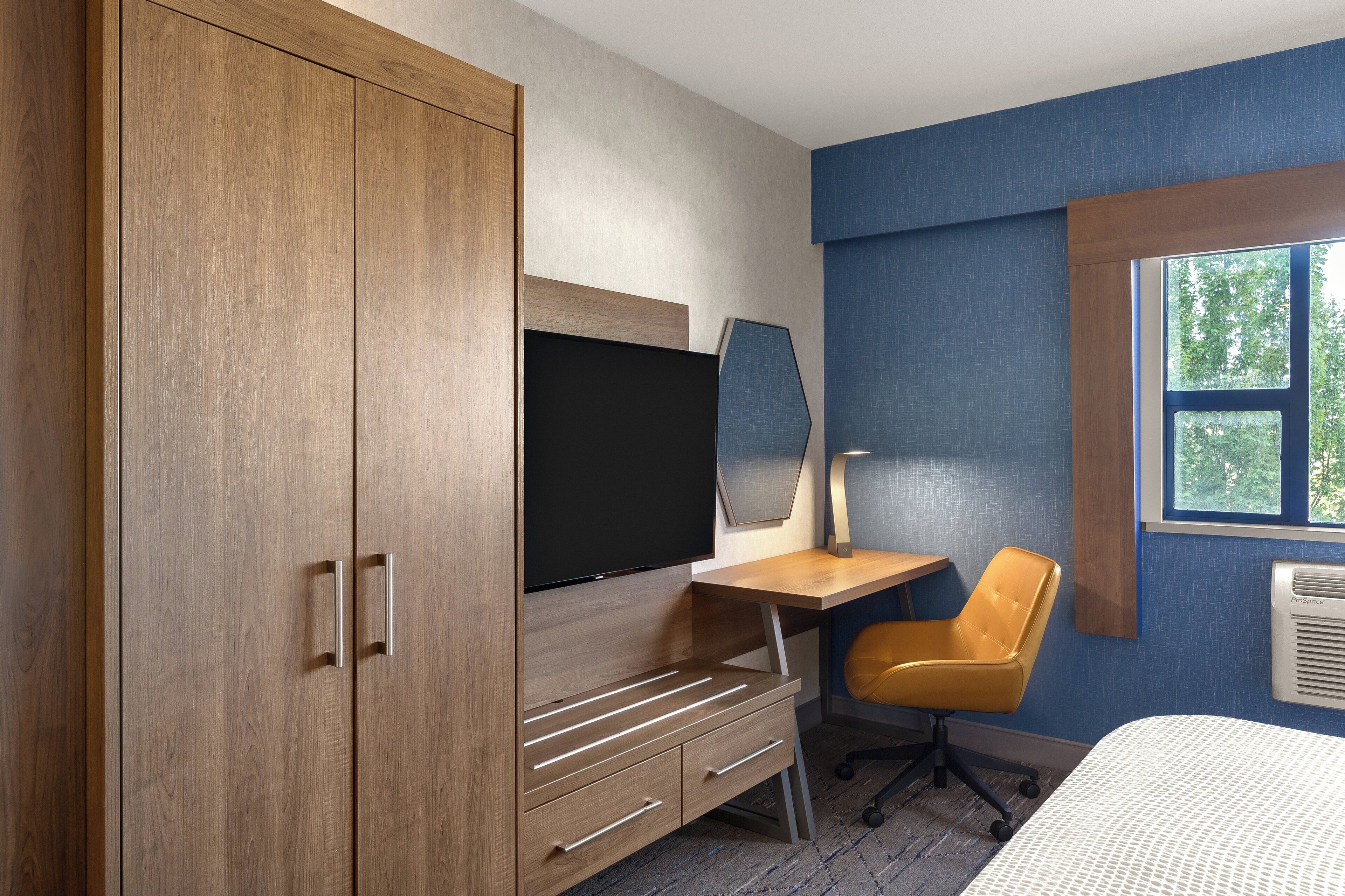Un dormitorio con un armario de madera, un escritorio con silla, una cama, un ventanal y un aire acondicionado de pared.
