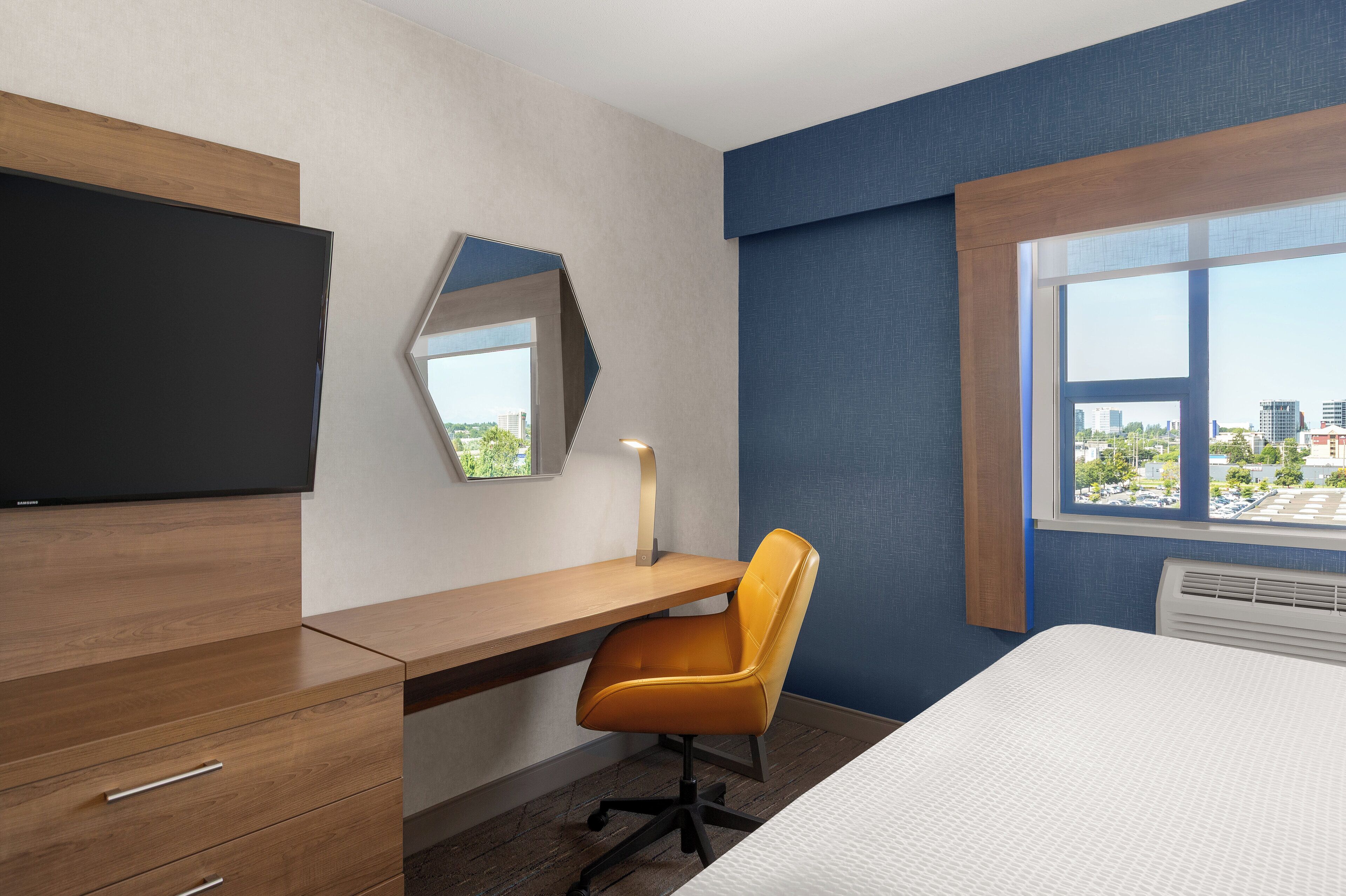 A hotel room with a bed, a desk, a chair, a TV, a window with a city view, and a mirror.