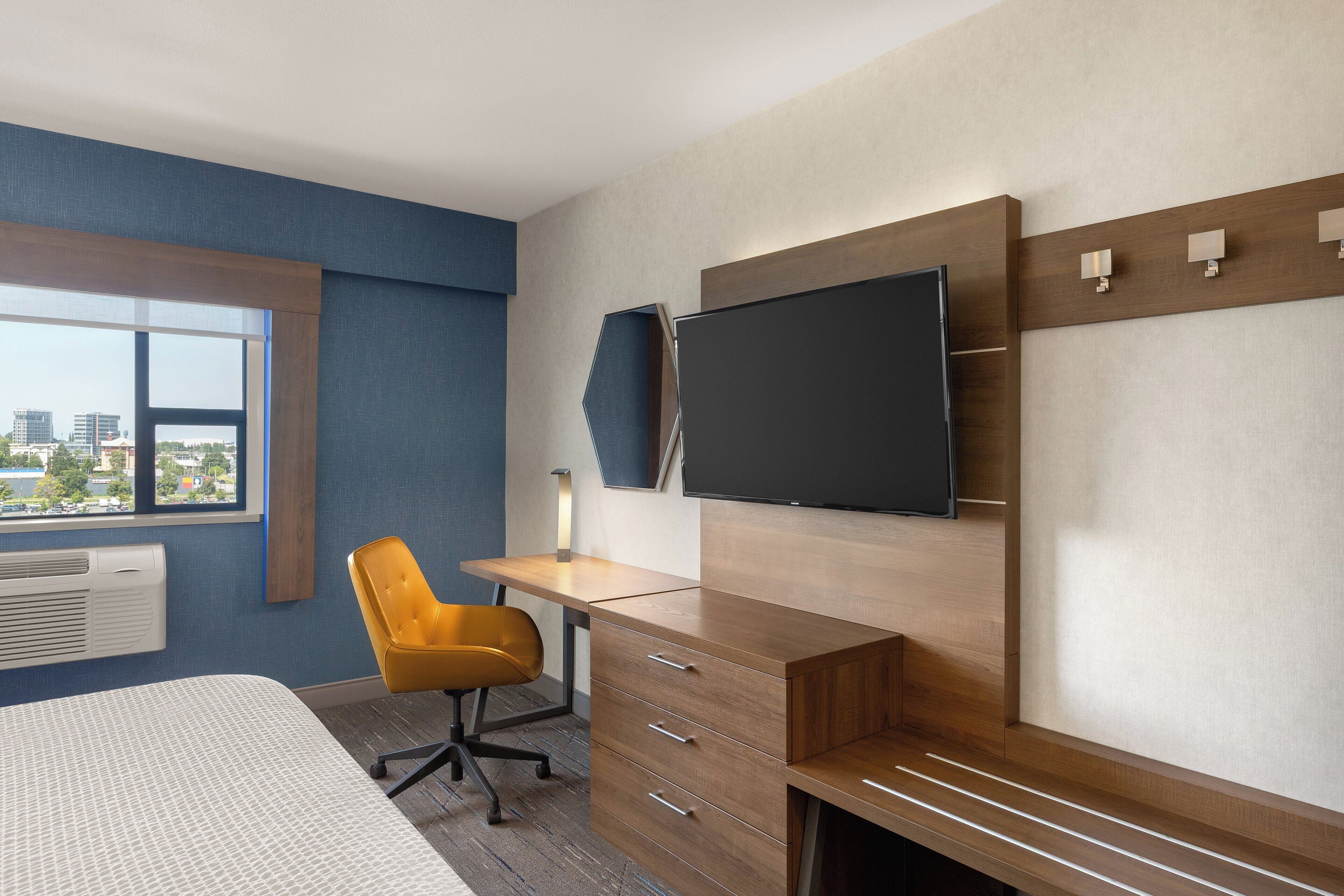 A hotel room with a bed, a desk with a chair, a TV mounted on the wall, and a window with a city view.
