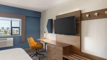 A hotel room with a bed, a desk with a chair, a TV mounted on the wall, and a window with a city view.