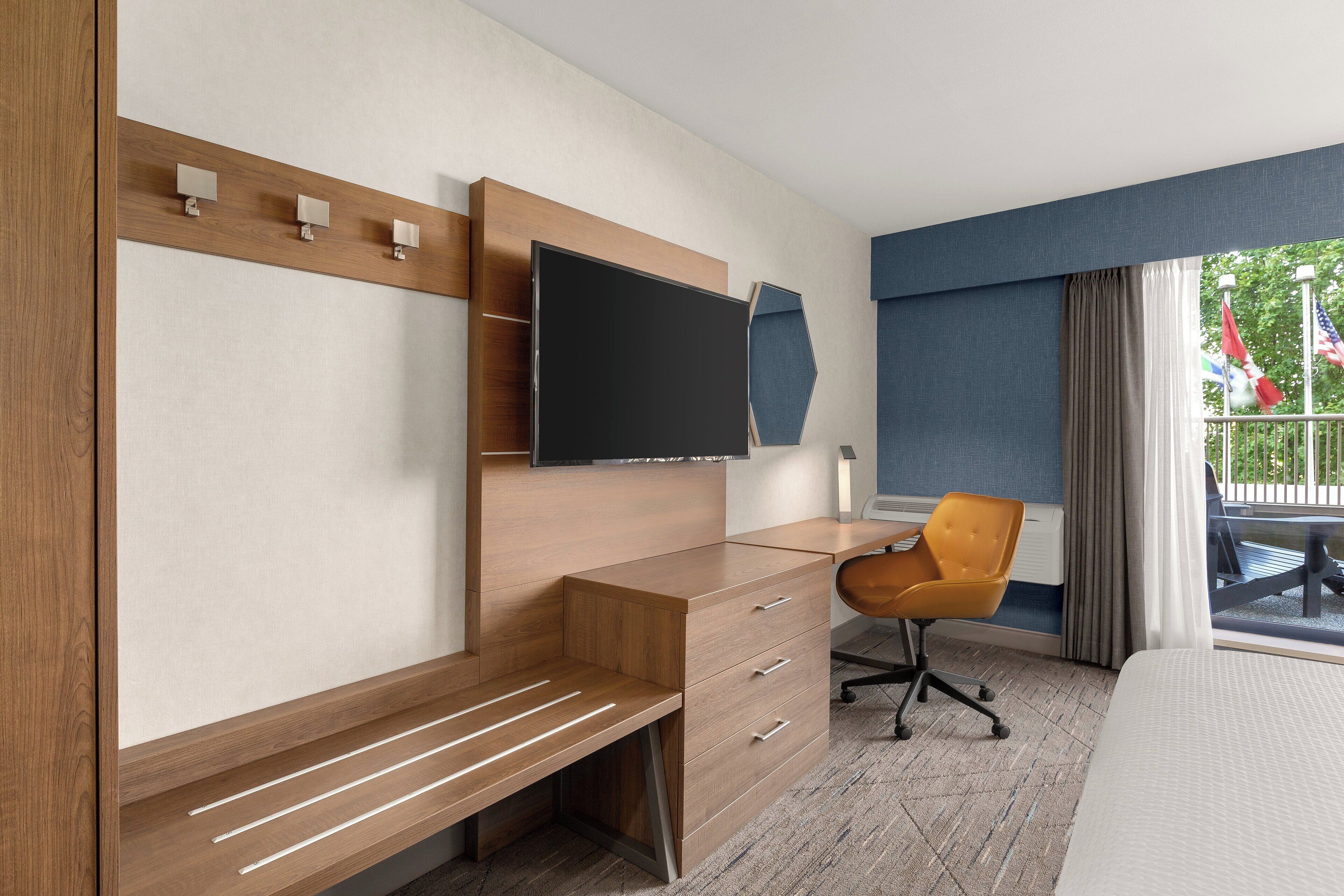 A hotel room with a wooden desk, a flat-screen TV, a comfortable chair, and a large window with a view of the outdoors.