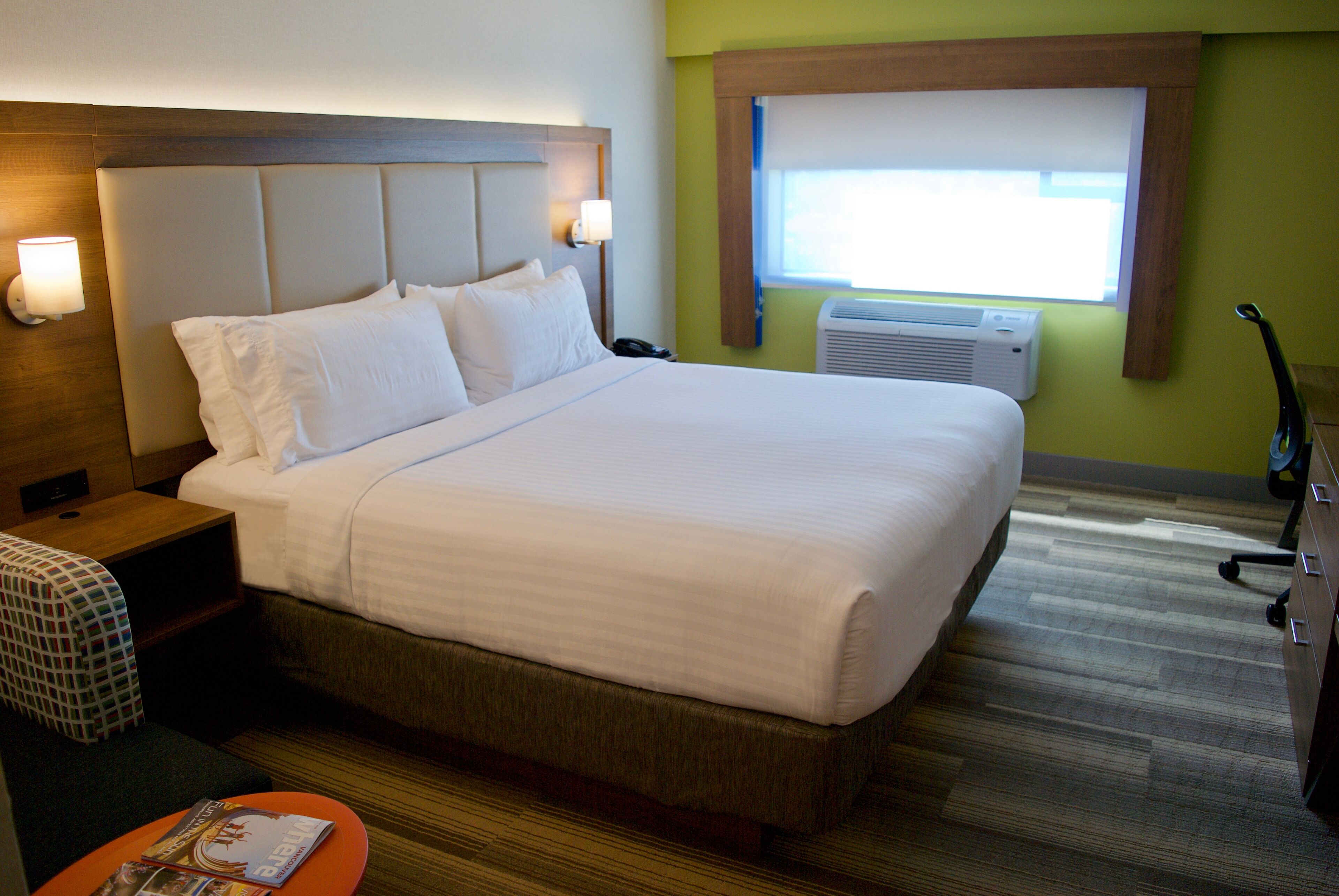 A hotel room with a large bed, bedside lamps, a window with an air conditioner, and a small table with a magazine.