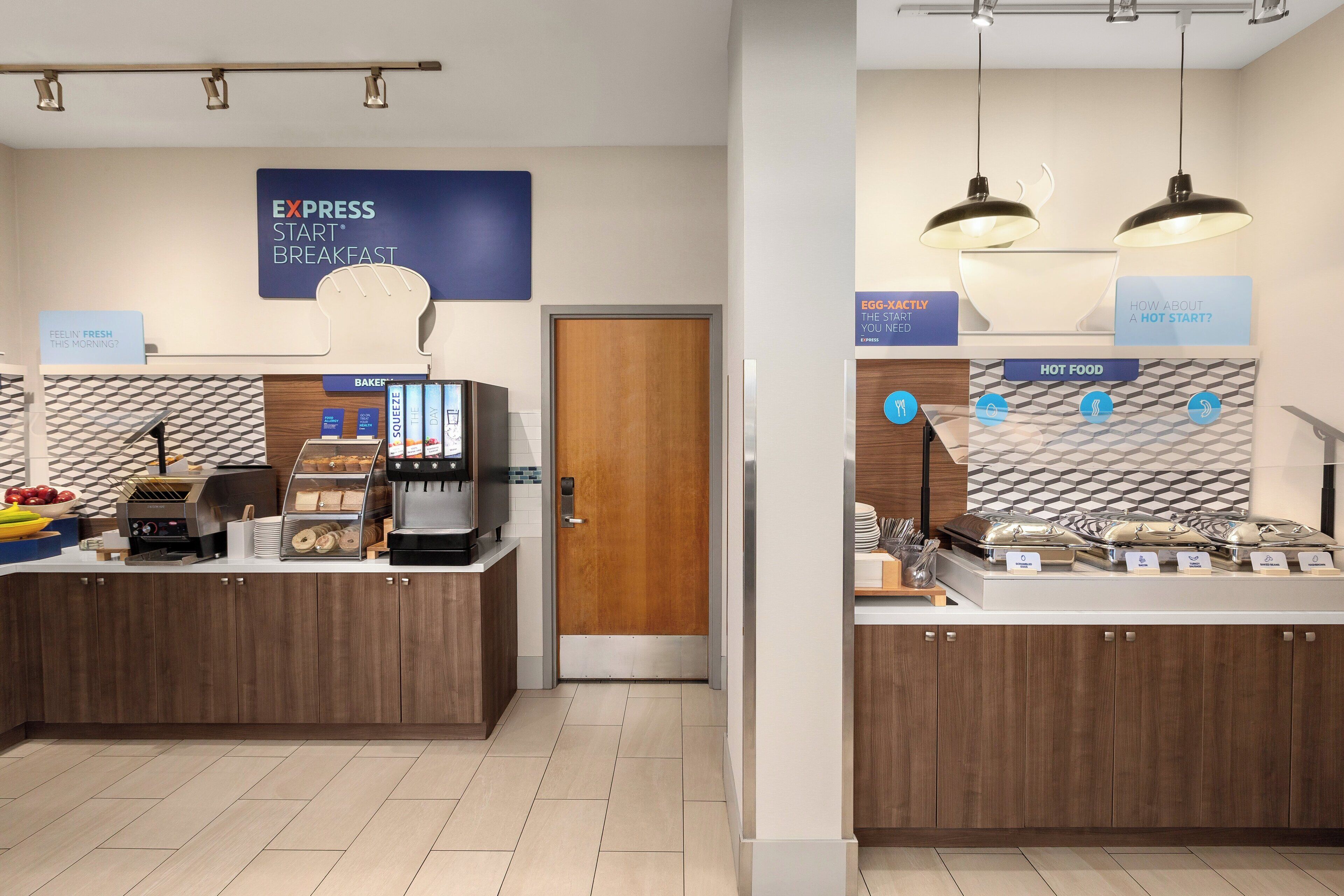 Espaço de refeições matinais com máquina de café, cesta de frutas e placa indicando Café da Manhã Express Start.