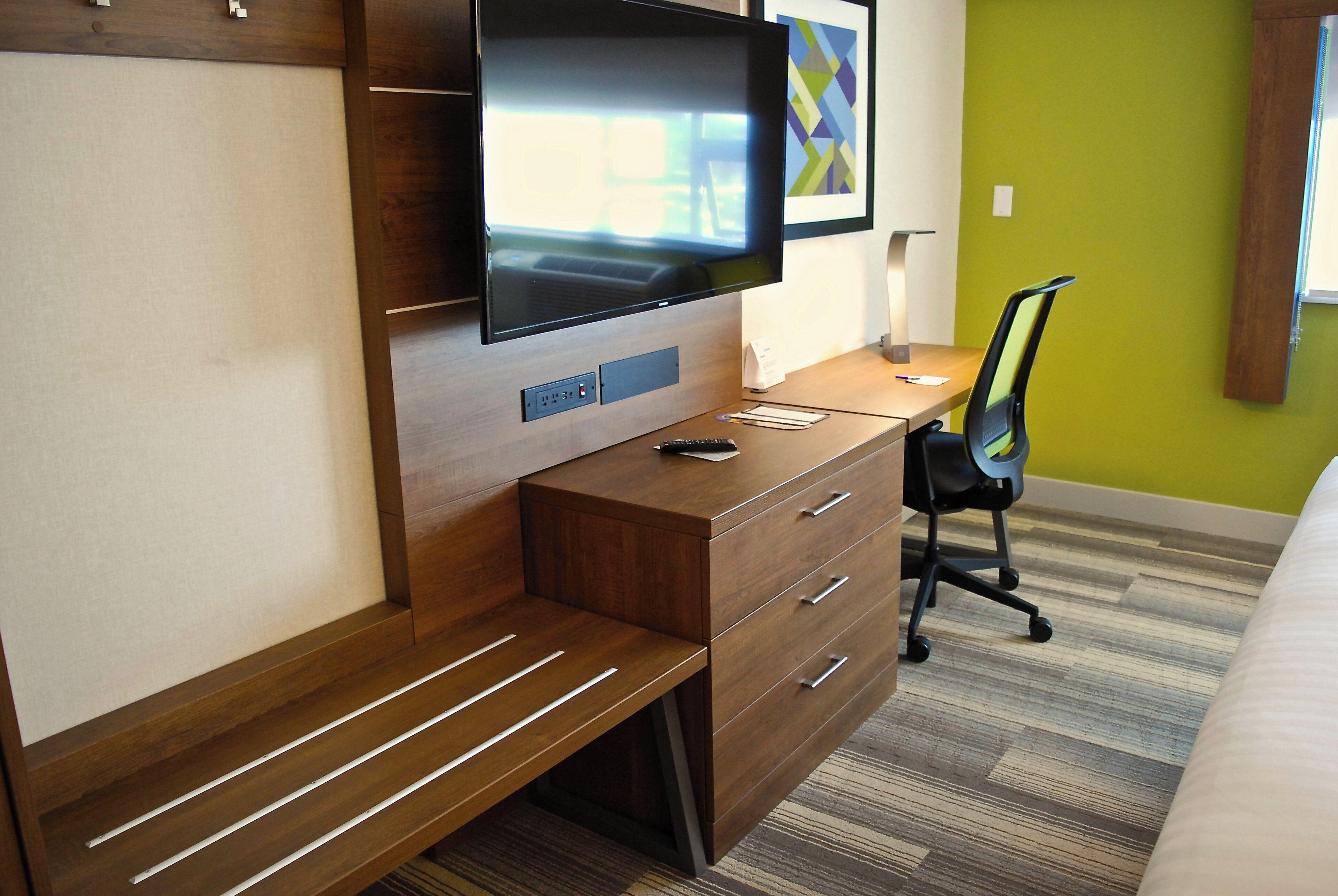 Une chambre d’hôtel avec un bureau en bois, un téléviseur à écran plat fixé au mur, un fauteuil de bureau noir et un lit recouvert d’un tapis à rayures.