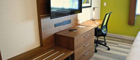 A hotel room with a wooden desk, a flat-screen TV mounted on the wall, a black office chair, and a bed with a striped carpet.