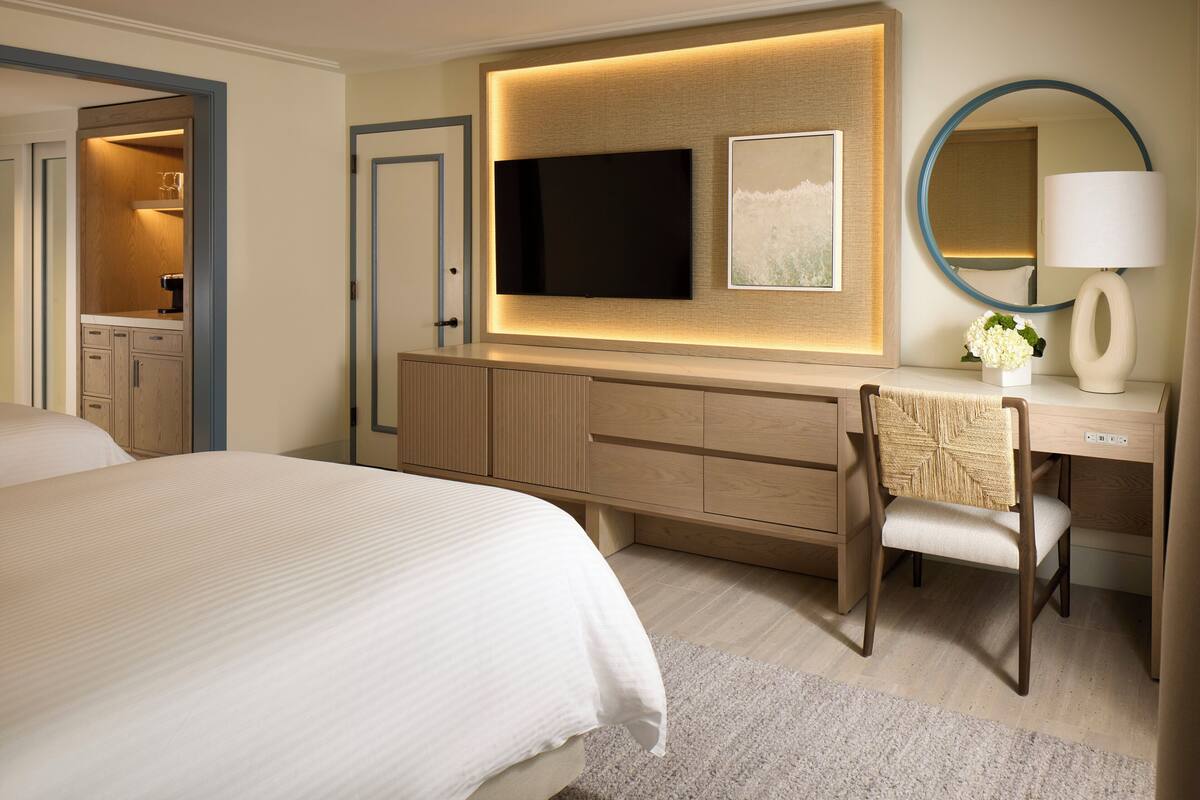 Une chambre d’hôtel avec un grand lit, un bureau avec une chaise, un téléviseur à écran plat, un miroir et une petite table sur laquelle est posé un vase de fleurs.