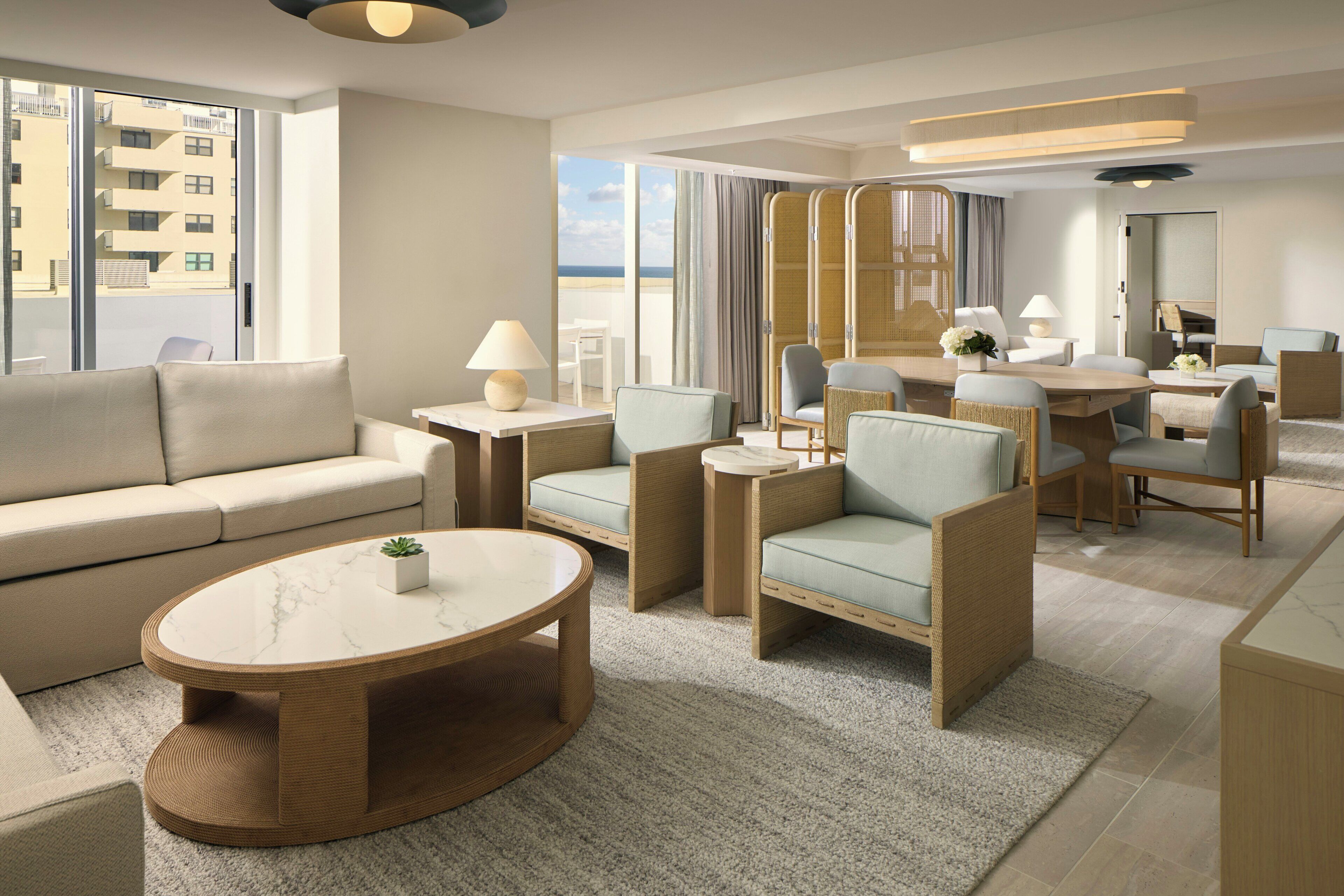 A modern hotel room with a sofa, armchairs, a dining table, and a view of the cityscape.