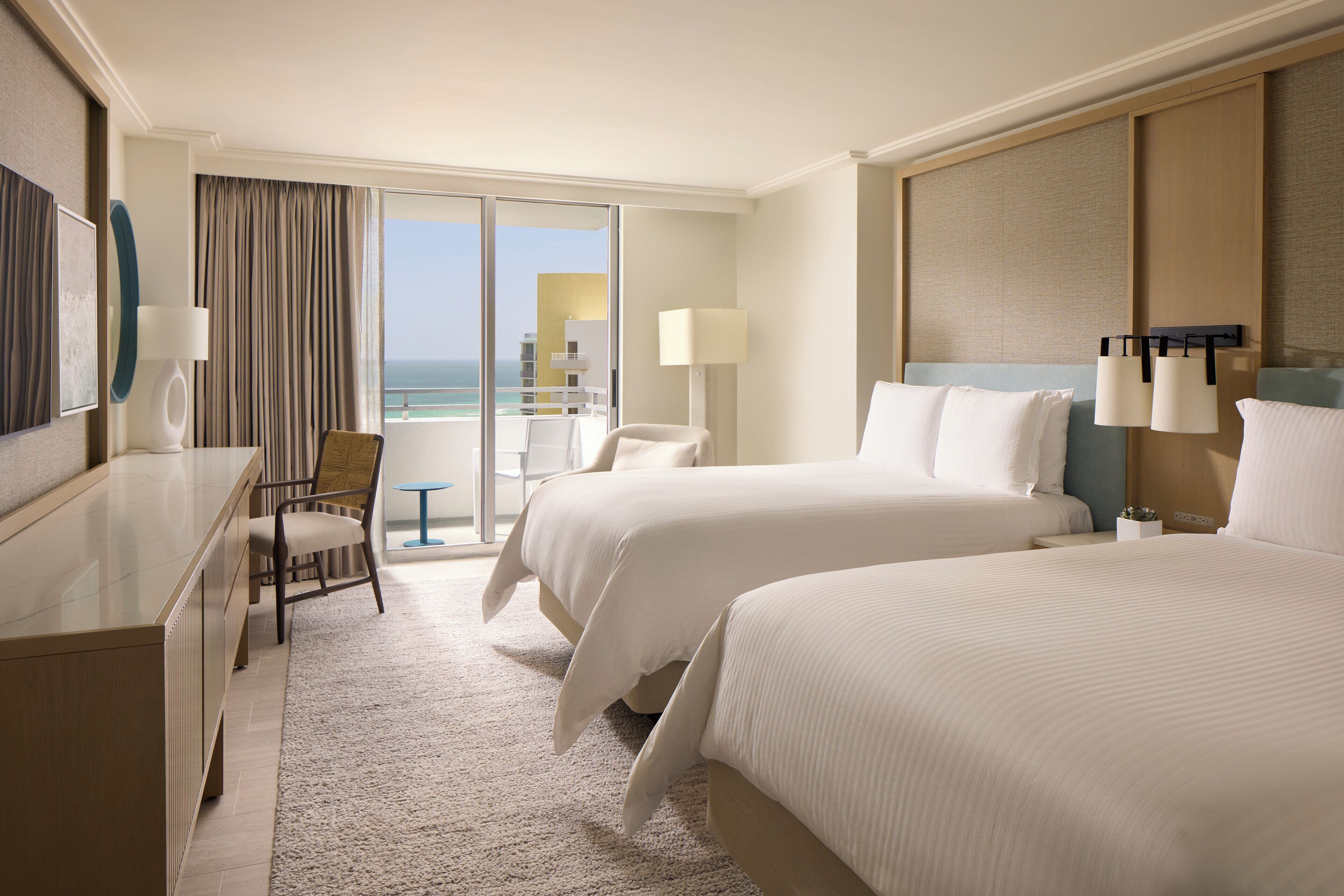 A hotel room with two beds, a desk, a chair, a lamp, and a view of the ocean.