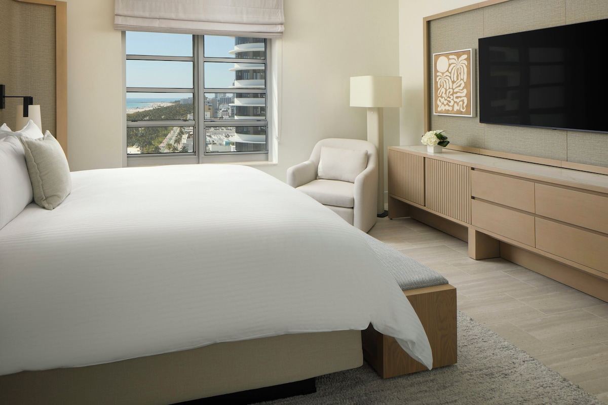 A modern hotel room with a large bed, a seating area, a TV, and a view of the city.