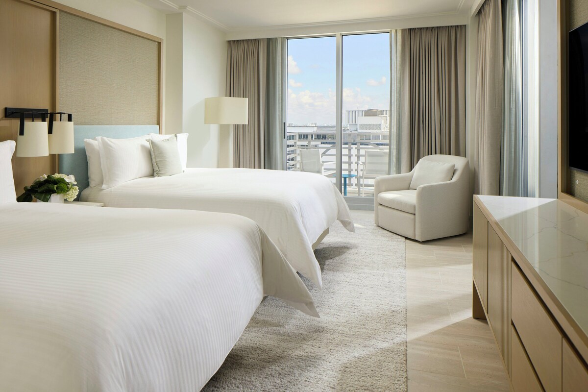 Une chambre d’hôtel avec deux lits, un bureau, une chaise, une télévision et une grande fenêtre avec des rideaux.