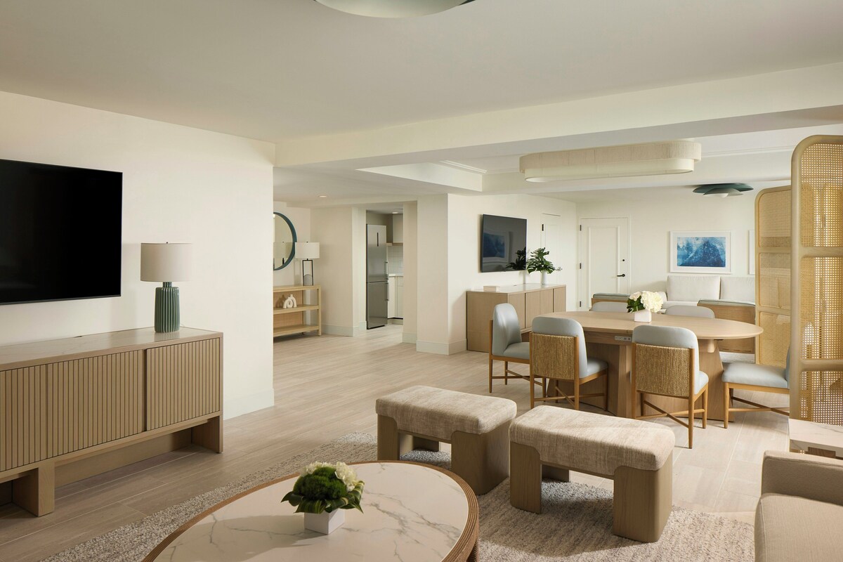 Una sala de estar moderna con un televisor de pantalla plana, una zona de comedor con una mesa redonda y sillas, y una cocina al fondo.