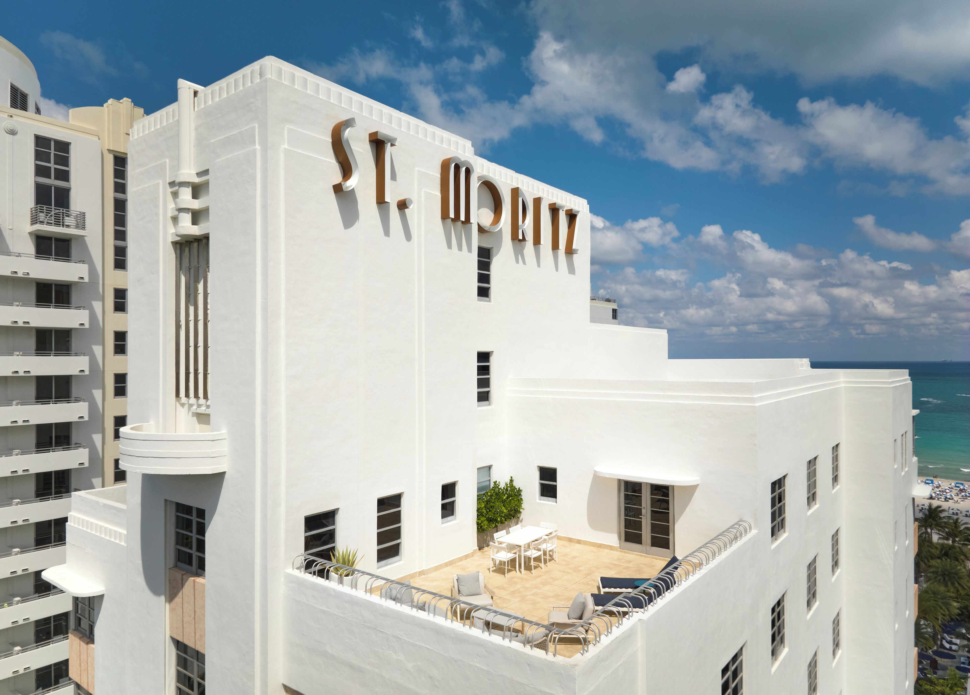 A white building with 'St. Moritz' on the facade, a rooftop patio with outdoor furniture, and a view of the ocean.