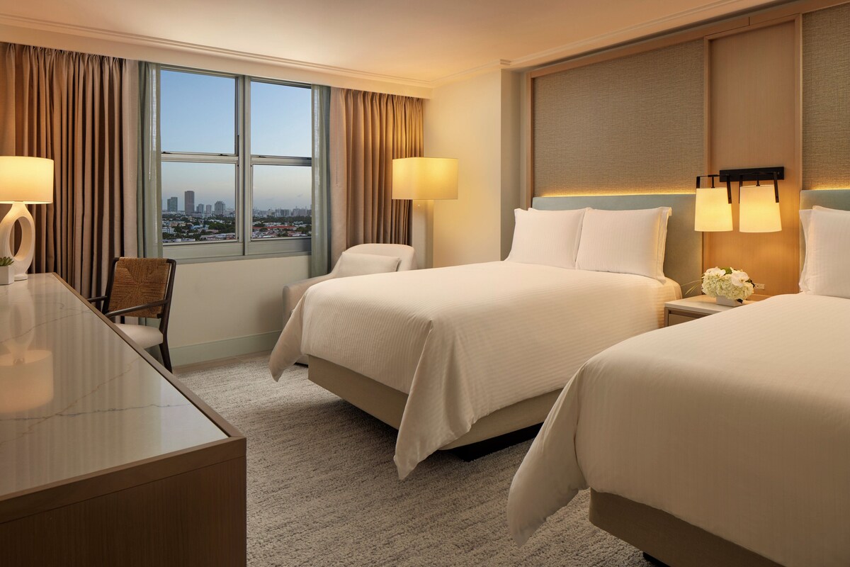 Habitación de hotel con dos camas, un escritorio y una silla. Hay dos lámparas en el escritorio y vistas de la ciudad por la ventana.
