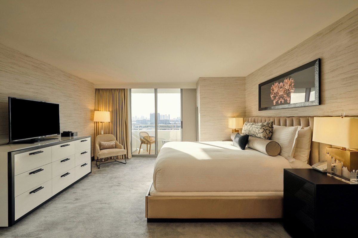 A modern hotel room with a large bed, a flat-screen TV, a desk, and a view of the city.