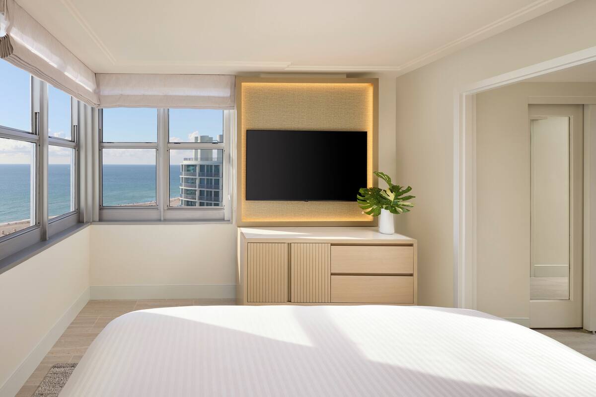 Une chambre d’hôtel avec un grand lit, un téléviseur à écran plat fixé au mur, une plante en pot et une vue sur l’océan par de grandes fenêtres.