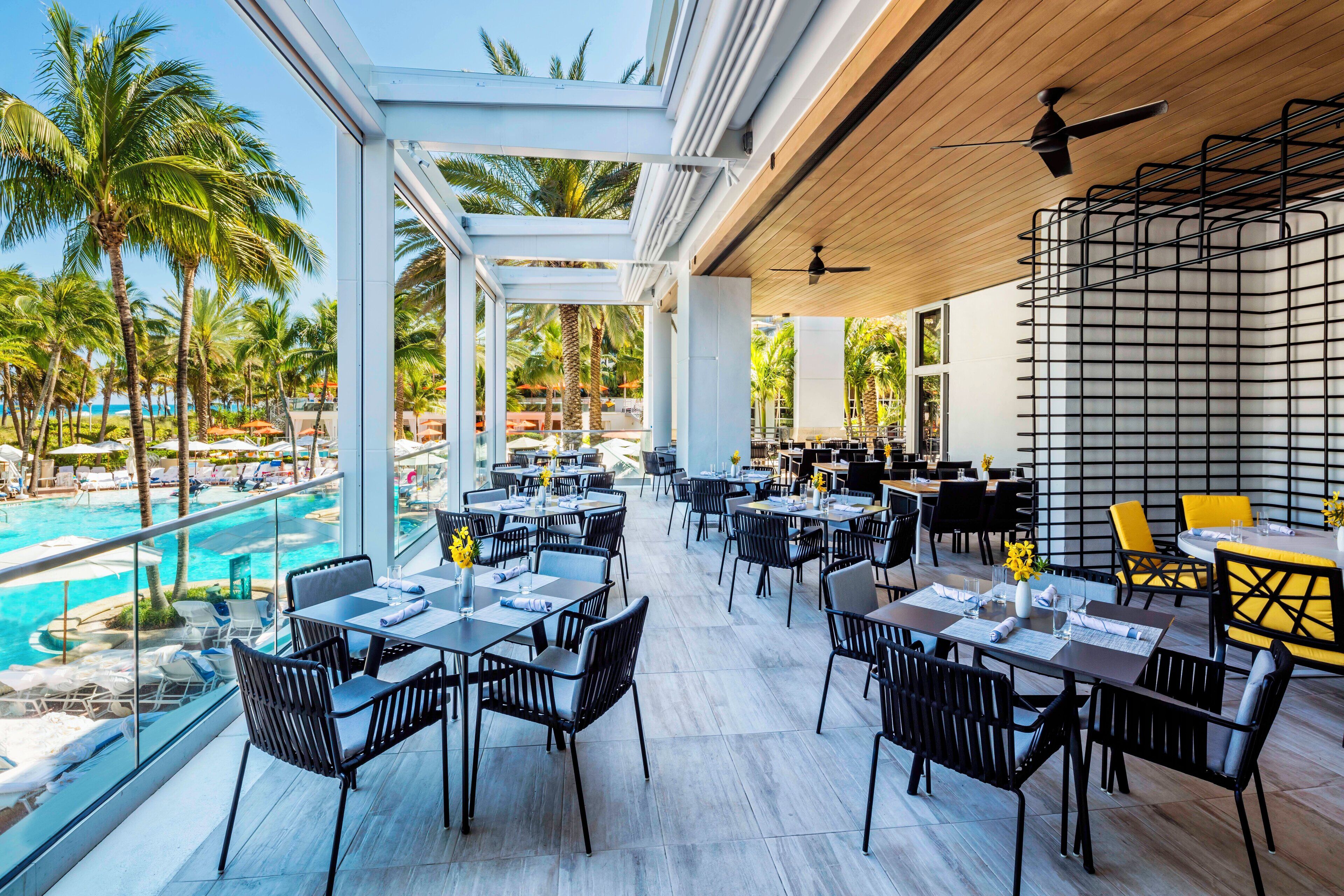 A rooftop dining area with tables and chairs, overlooking a pool and palm trees.